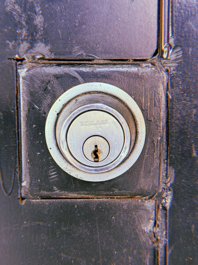 Silver Schlage deadbolt lock mounted on a dark painted door, surrounded by a square recessed panel with scratched paint and worn edges.