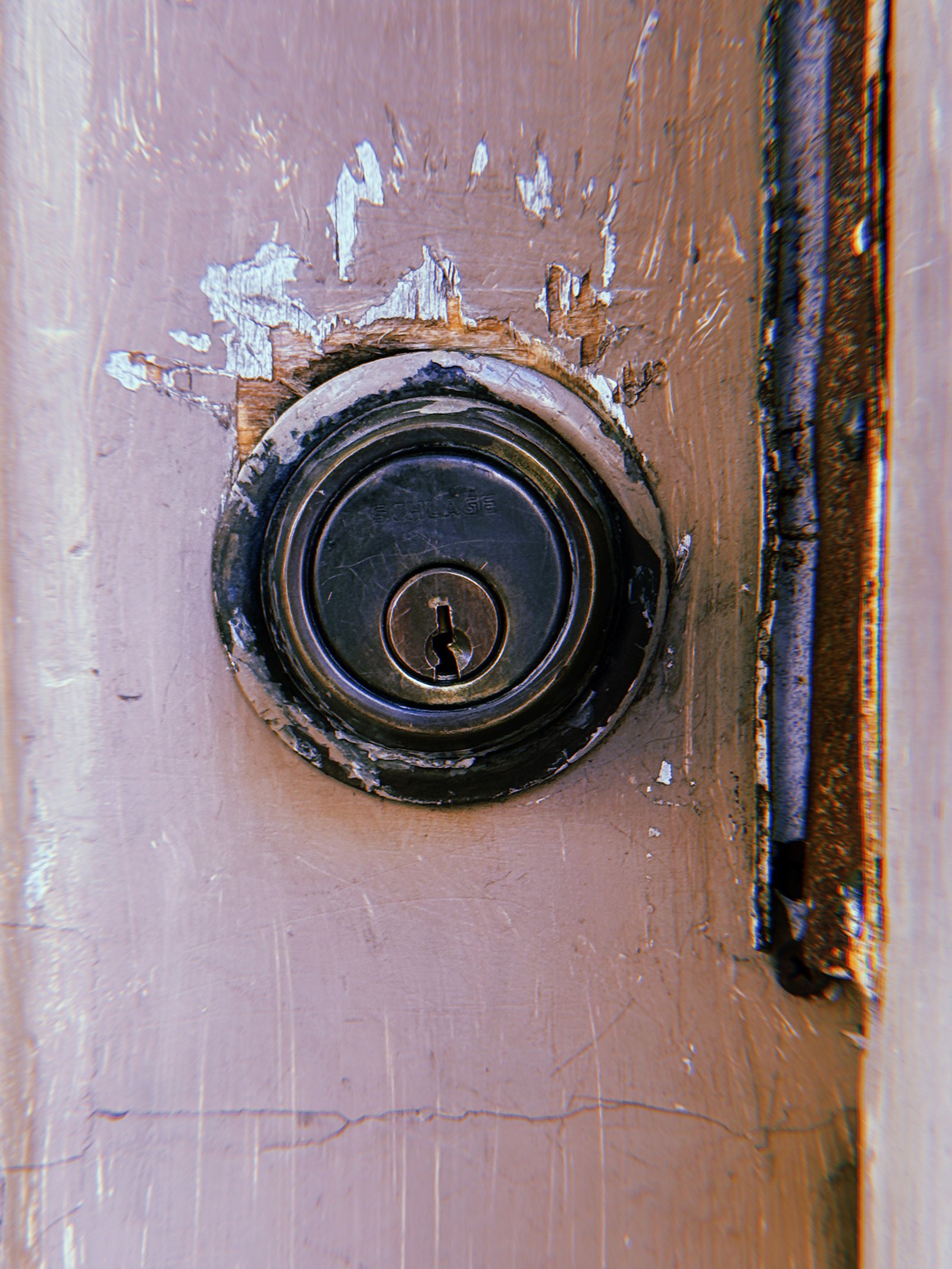 Weathered Schlage deadbolt lock mounted on a pale pink wooden door, surrounded by peeling paint, exposed wood, and scratched surfaces.