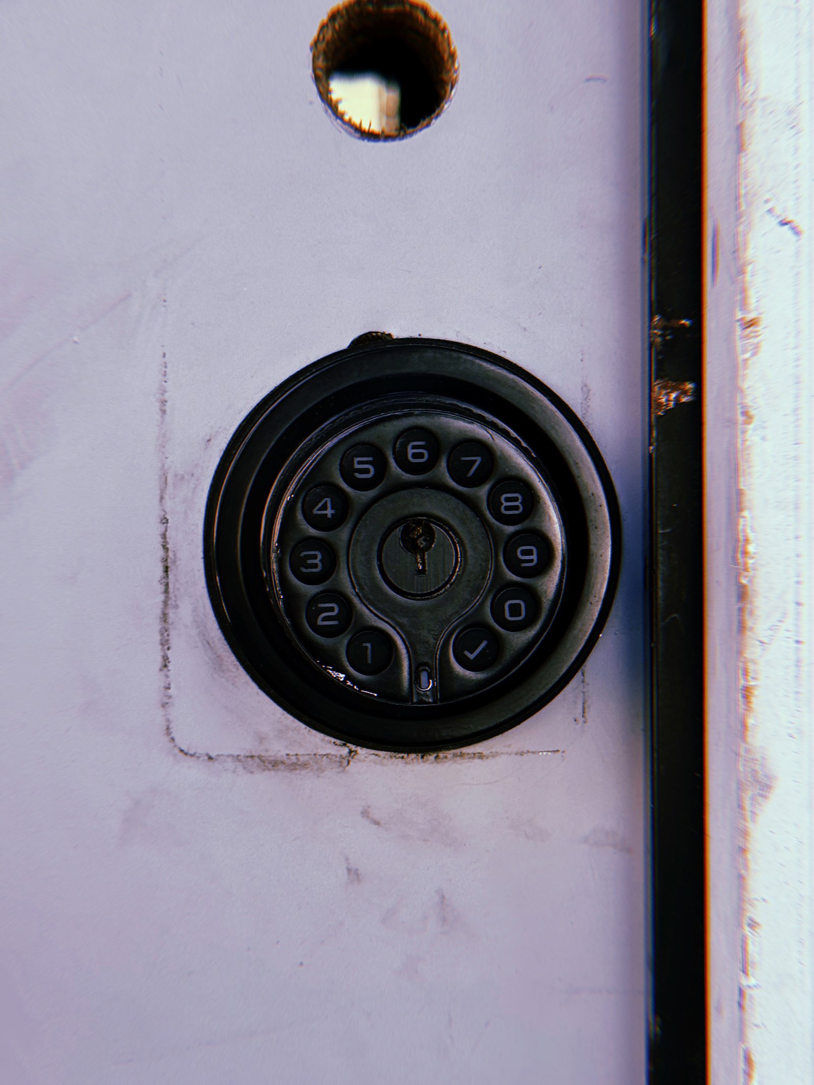 Black circular electronic keypad lock with numbered buttons and a central keyhole mounted on a pale lavender metal door, beneath an empty circular opening.