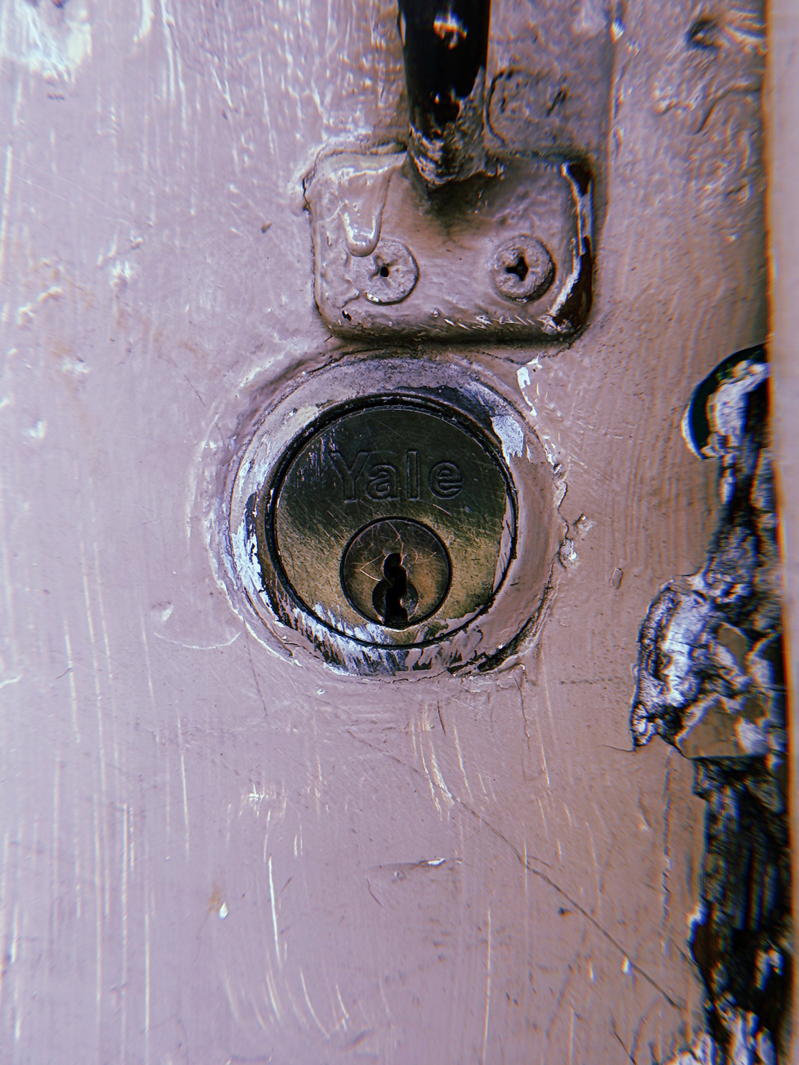 Weathered Yale cylinder lock embedded in a scratched, pale pink painted metal door, with chipped paint around the keyhole and visible mounting hardware above.