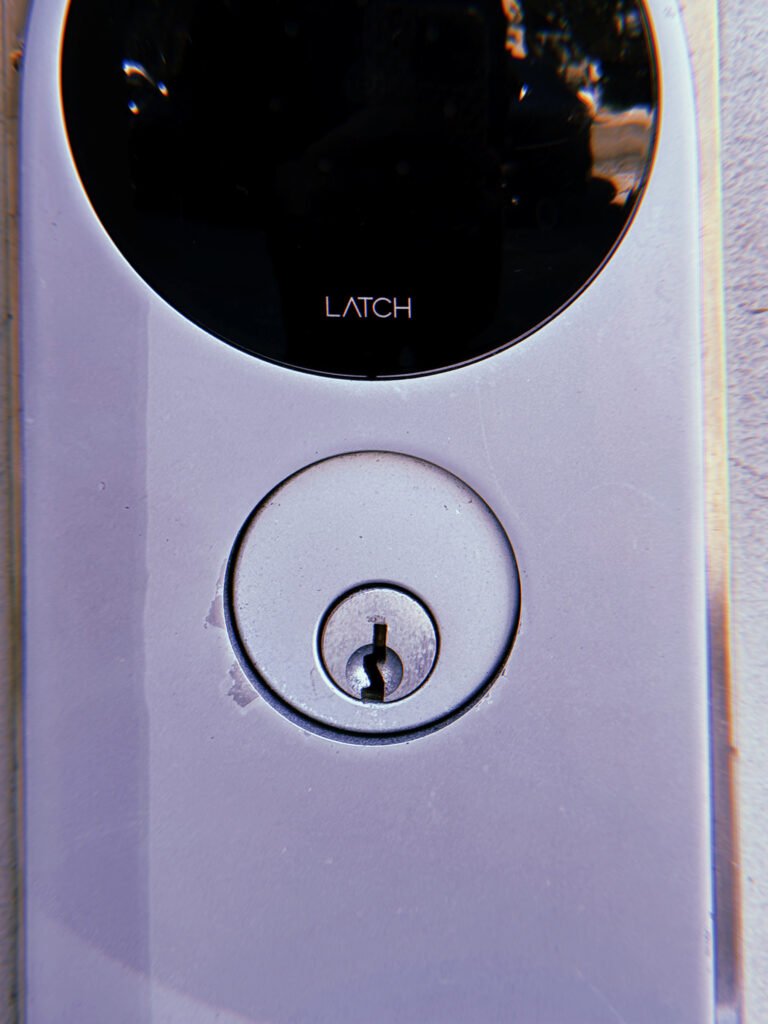 Silver circular keyhole lock mounted beneath a black LATCH smart lock panel on a light metallic door surface.