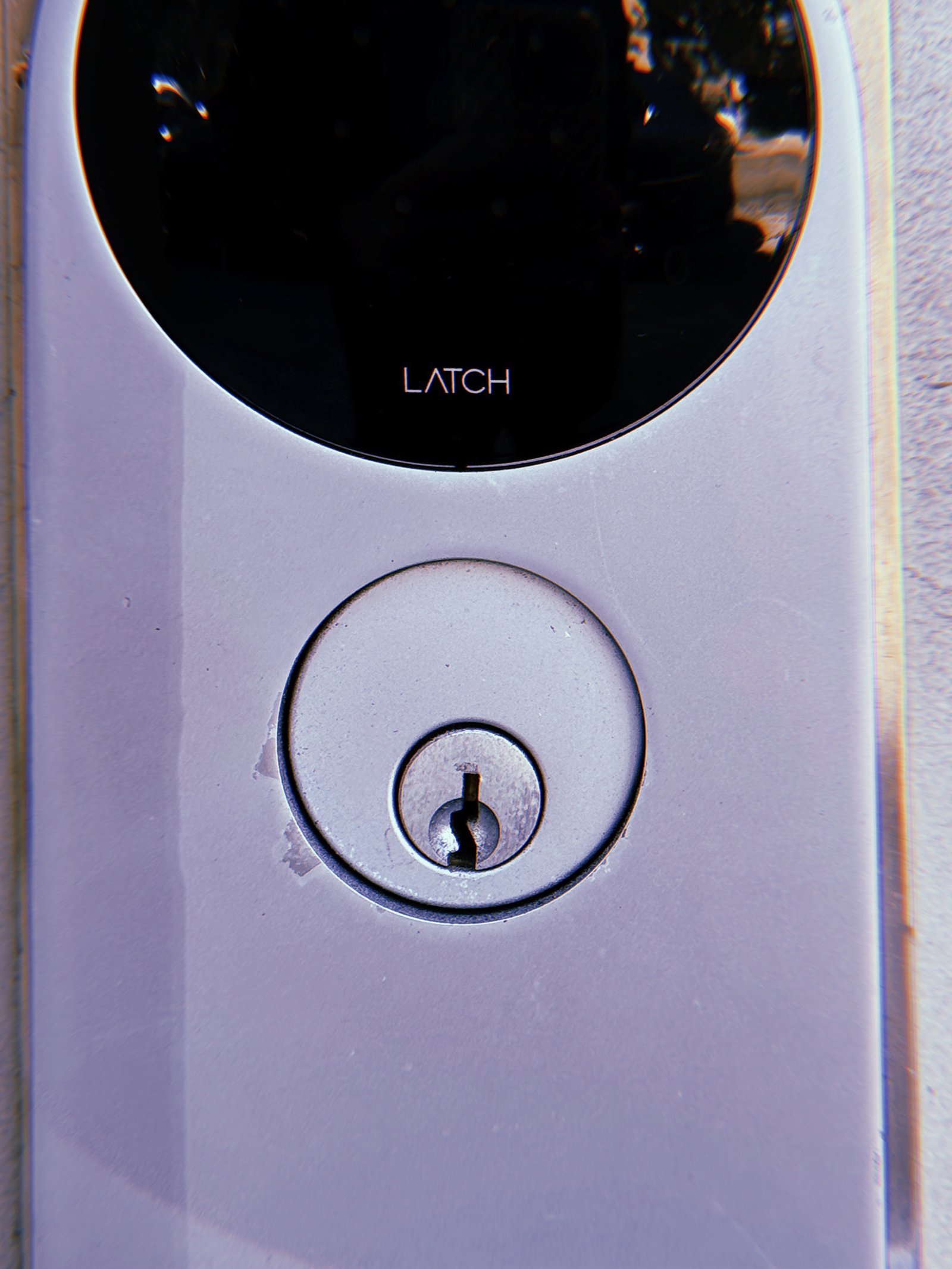 Silver circular keyhole lock mounted beneath a black LATCH smart lock panel on a light metallic door surface.