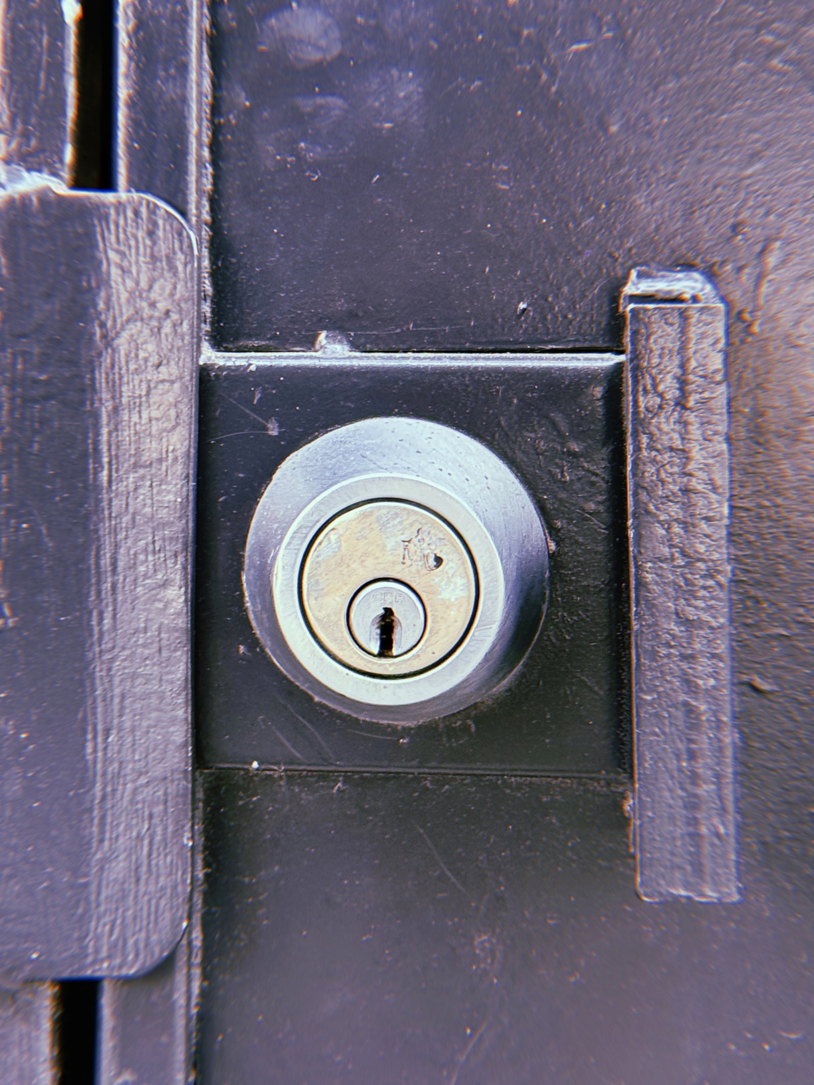 Silver circular door lock cylinder mounted on a dark metal door, positioned between two vertical metal strips with worn paint and visible scratches.