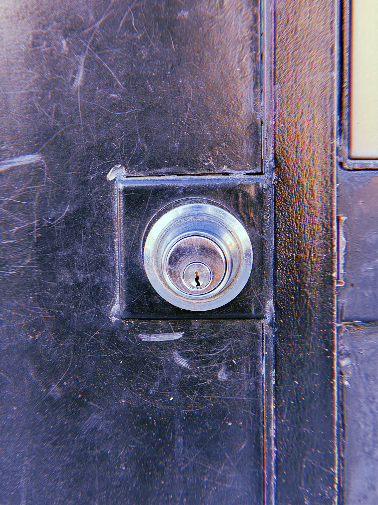 Silver circular door lock cylinder mounted on a square black metal plate, surrounded by scratches and worn paint on a dark door.