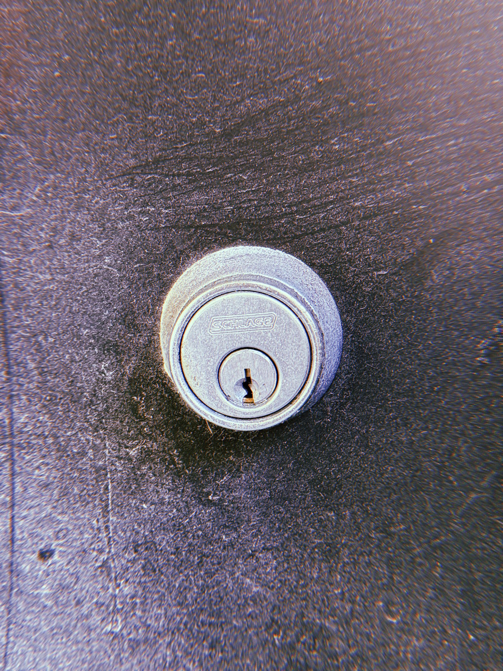 Close-up of a silver Schlage door lock cylinder mounted on a dark textured metal surface, with a central keyhole and circular metal housing.