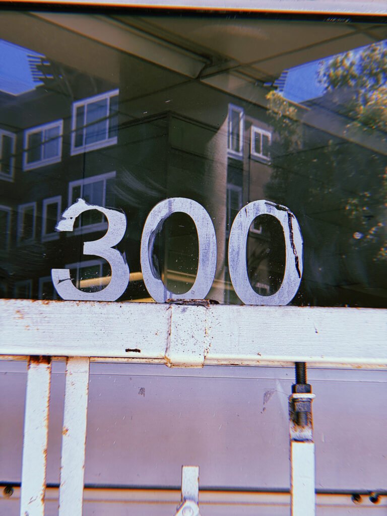 Weathered white address numbers 300 attached to a glass entrance, reflecting a residential building with multiple windows across the street.