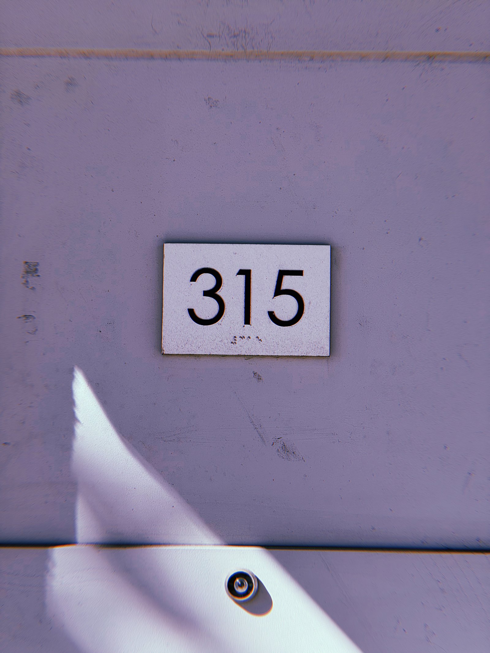 ALT Text: White rectangular address plaque displaying black numbers 315 on a pale lavender wall, with diagonal sunlight and a small circular doorbell below.