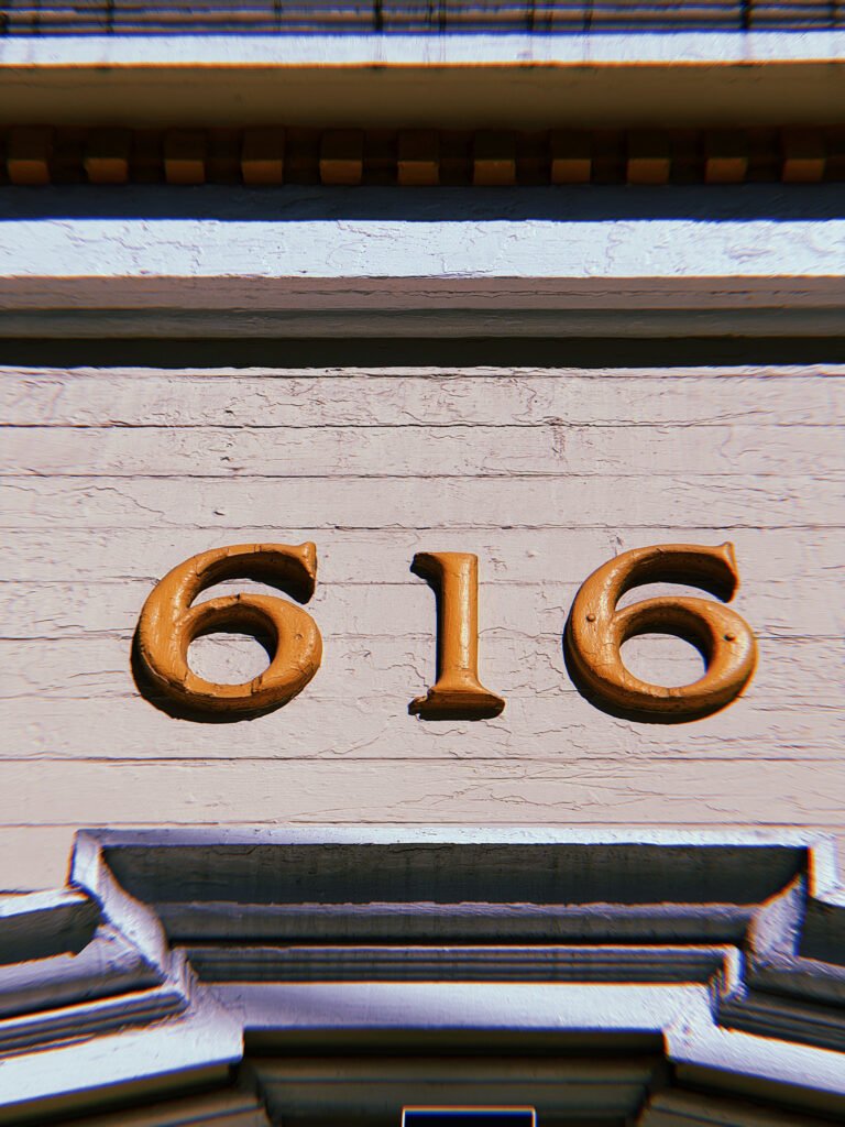 Gold address numbers 616 mounted horizontally on a weathered white wooden facade above an entrance with decorative architectural molding.