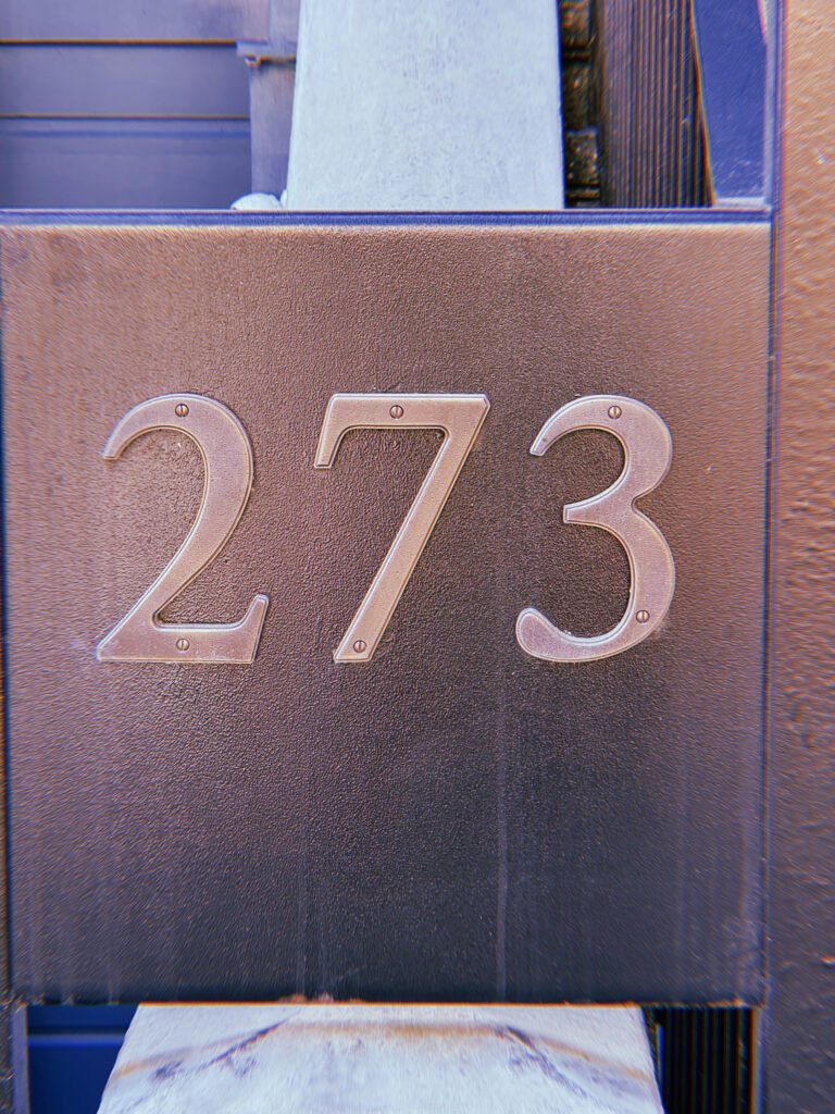 Metal address numbers 273 mounted horizontally on a dark brown textured rectangular panel attached to a building facade.