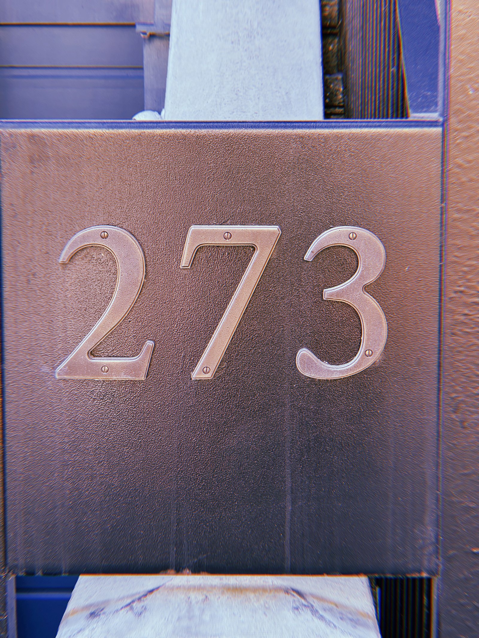 Metal address numbers 273 mounted horizontally on a dark brown textured rectangular panel attached to a building facade.