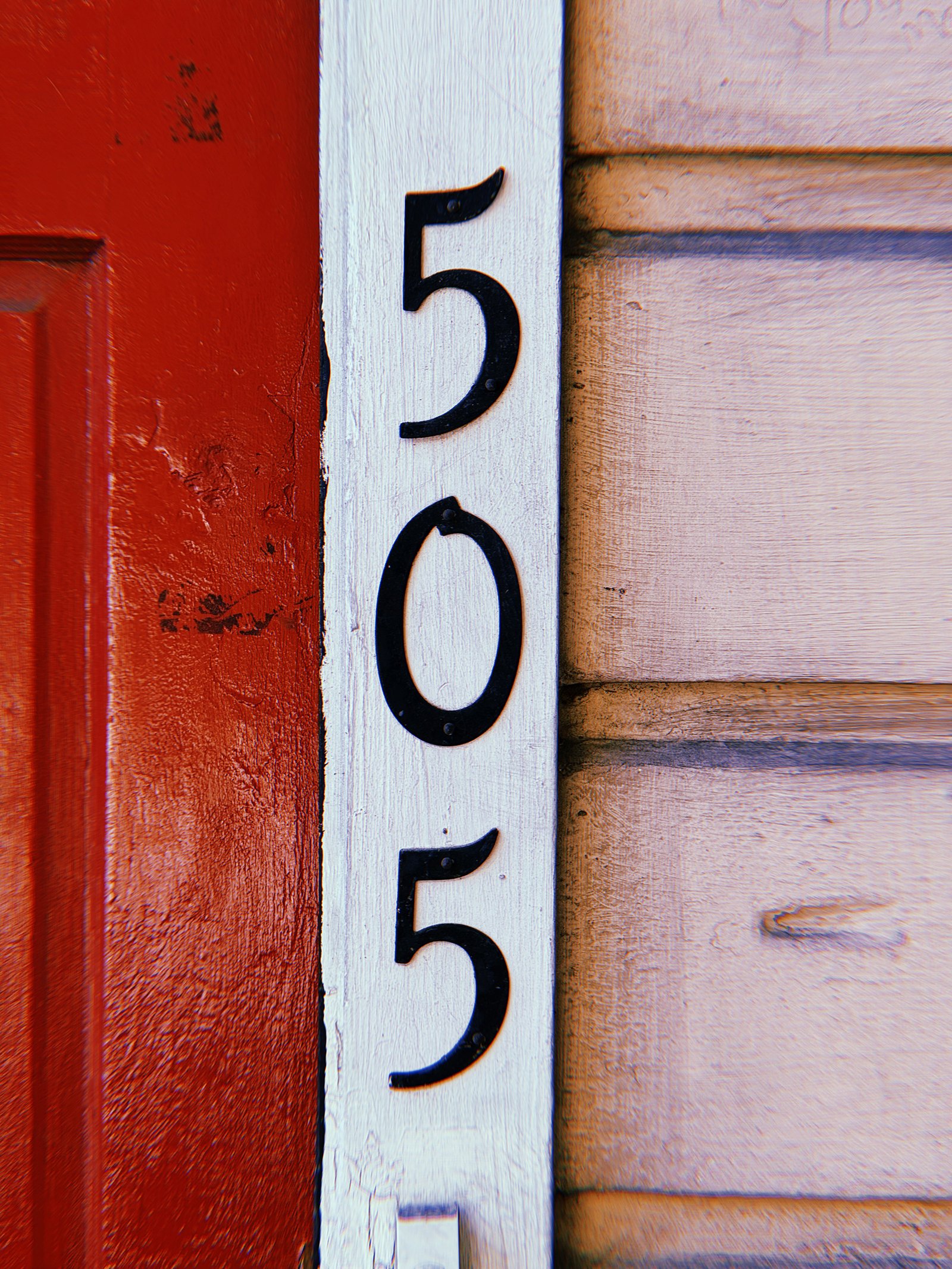 Black metal address numbers 505 arranged vertically on white wooden trim between a red door and beige horizontal siding.