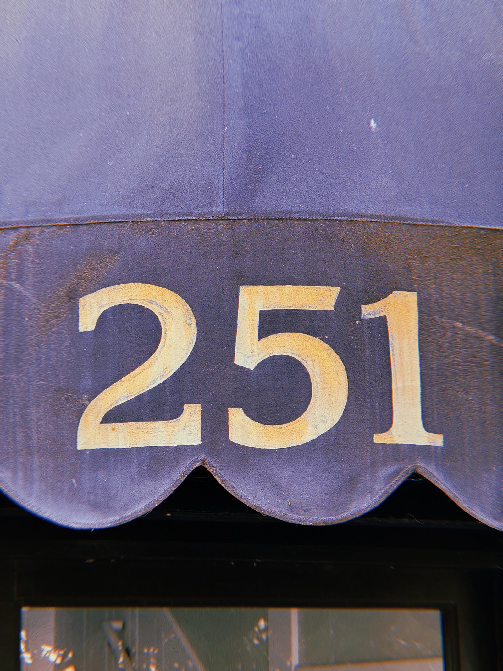 Gold address numbers 251 painted on a dark blue curved awning with a scalloped lower edge above a building entrance.