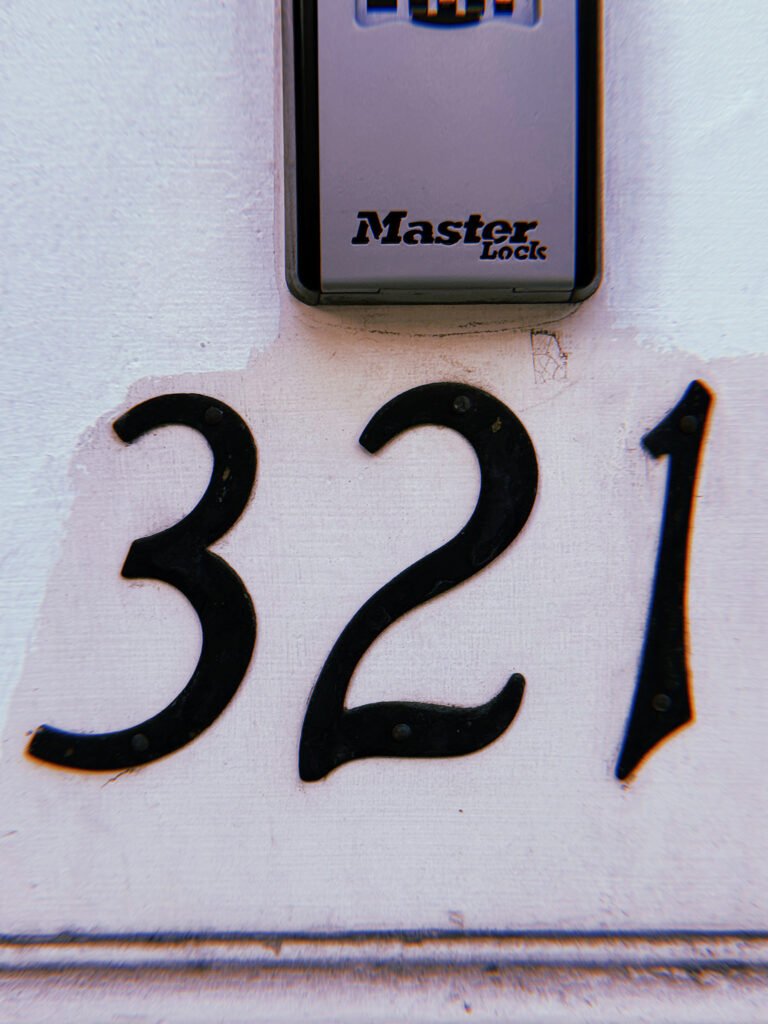 Black metal address numbers 321 mounted on a pale painted wall, with a gray Master Lock combination lockbox positioned above.