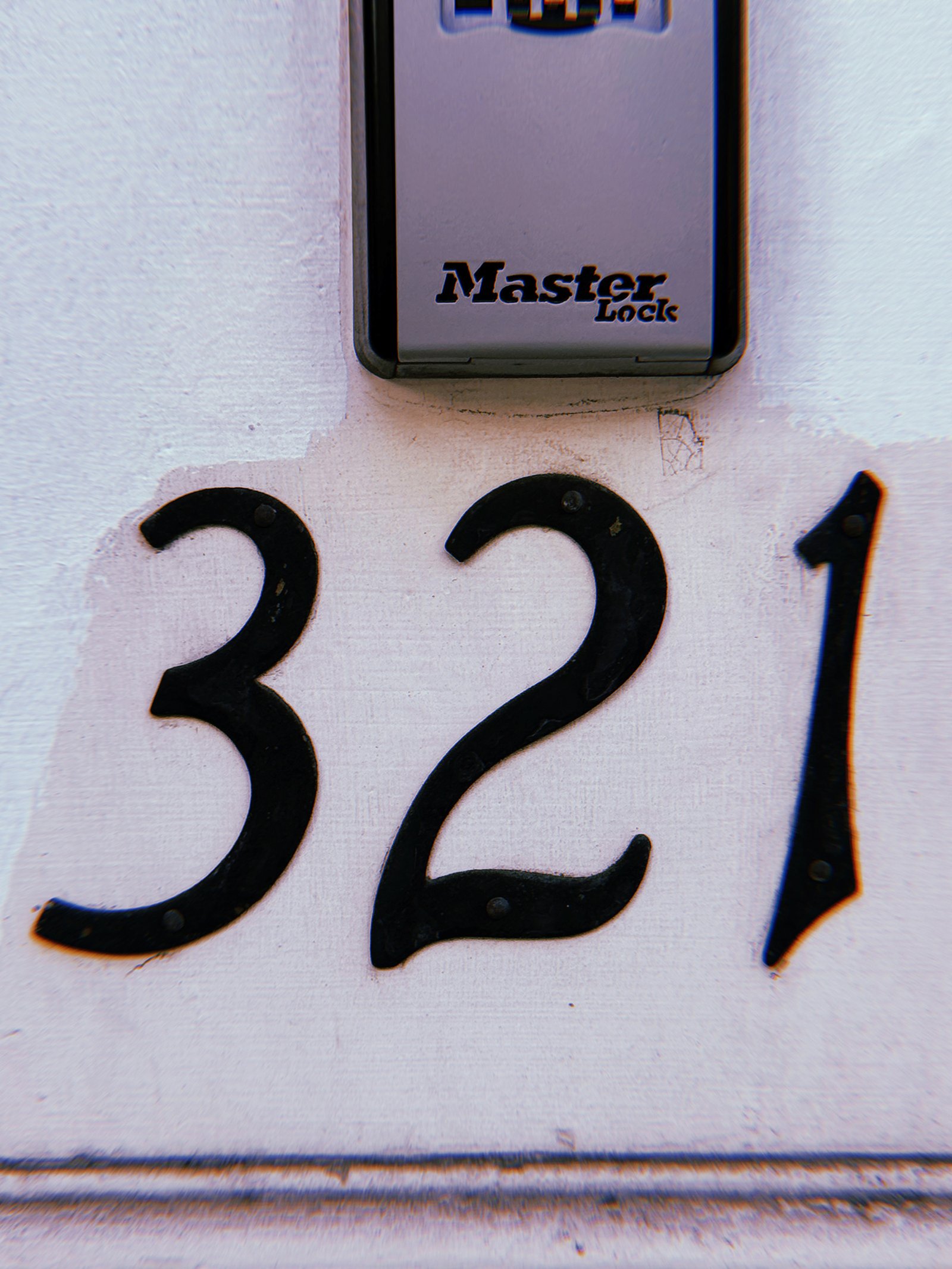 Black metal address numbers 321 mounted on a pale painted wall, with a gray Master Lock combination lockbox positioned above.