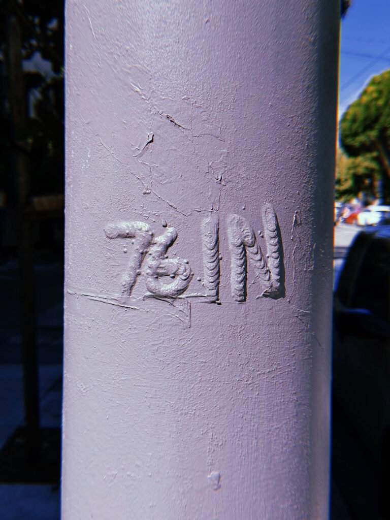 Raised "761 N" marking on a pale blue-gray metal utility pole in San Francisco. Thick layers of paint cover the embossed characters, with a blurred street and parked vehicles in the background.