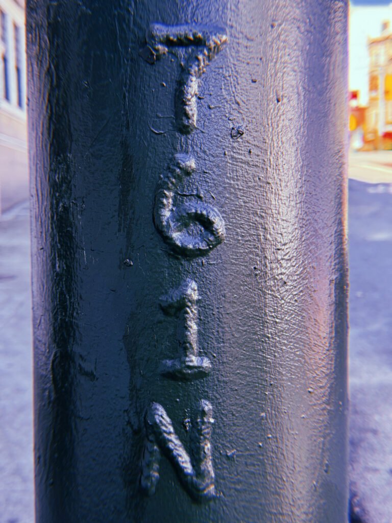 Vertical "761 N" marking embossed on a dark blue-gray metal utility pole in San Francisco. Thick layers of paint cover the raised characters, with a blurred street intersection in the background.