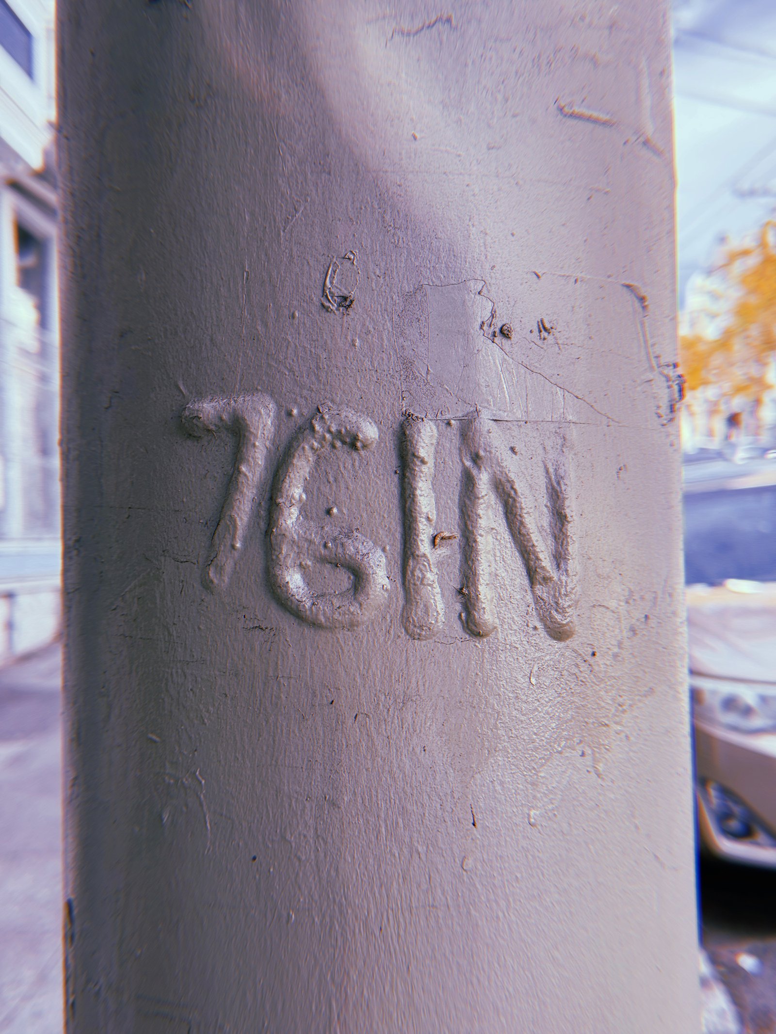 Raised "76 IN" marking embossed on a gray-painted metal utility pole in San Francisco. Thick layers of paint partially obscure the characters, with scratches and a city street visible in the background.
