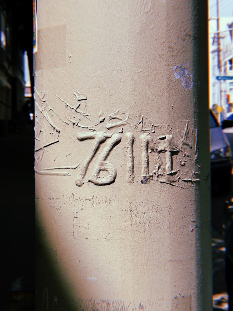 Raised "76 IE" marking on a beige-painted metal utility pole in San Francisco. Thick, cracked layers of paint partially obscure the embossed characters, with a city street visible in the background.