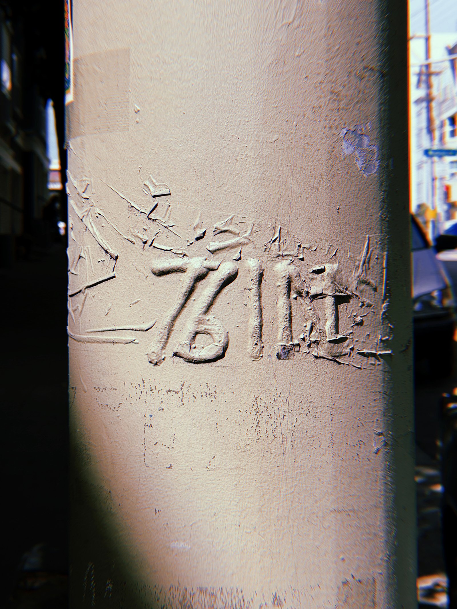 Raised "76 IE" marking on a beige-painted metal utility pole in San Francisco. Thick, cracked layers of paint partially obscure the embossed characters, with a city street visible in the background.