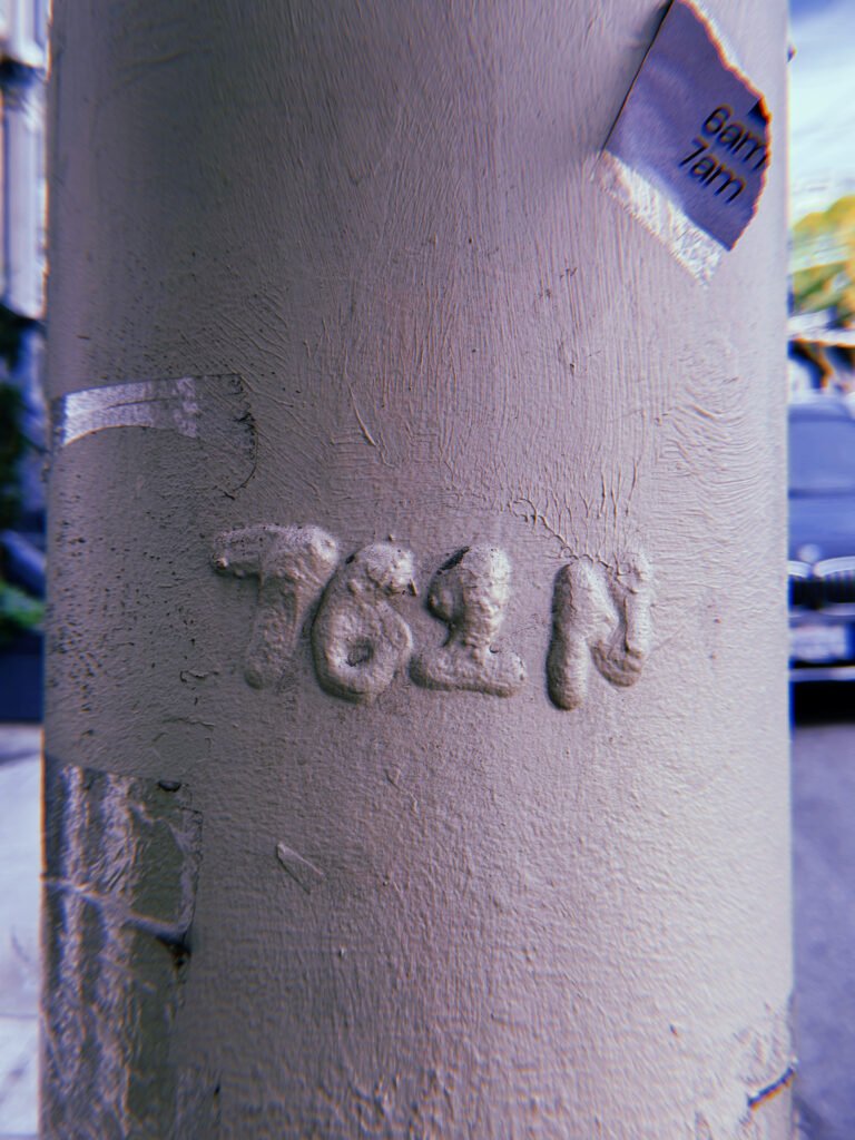 Raised "762 N" marking embossed on a gray-painted metal utility pole in San Francisco. Thick layers of paint partially obscure the characters, with remnants of stickers and surface wear visible around them.