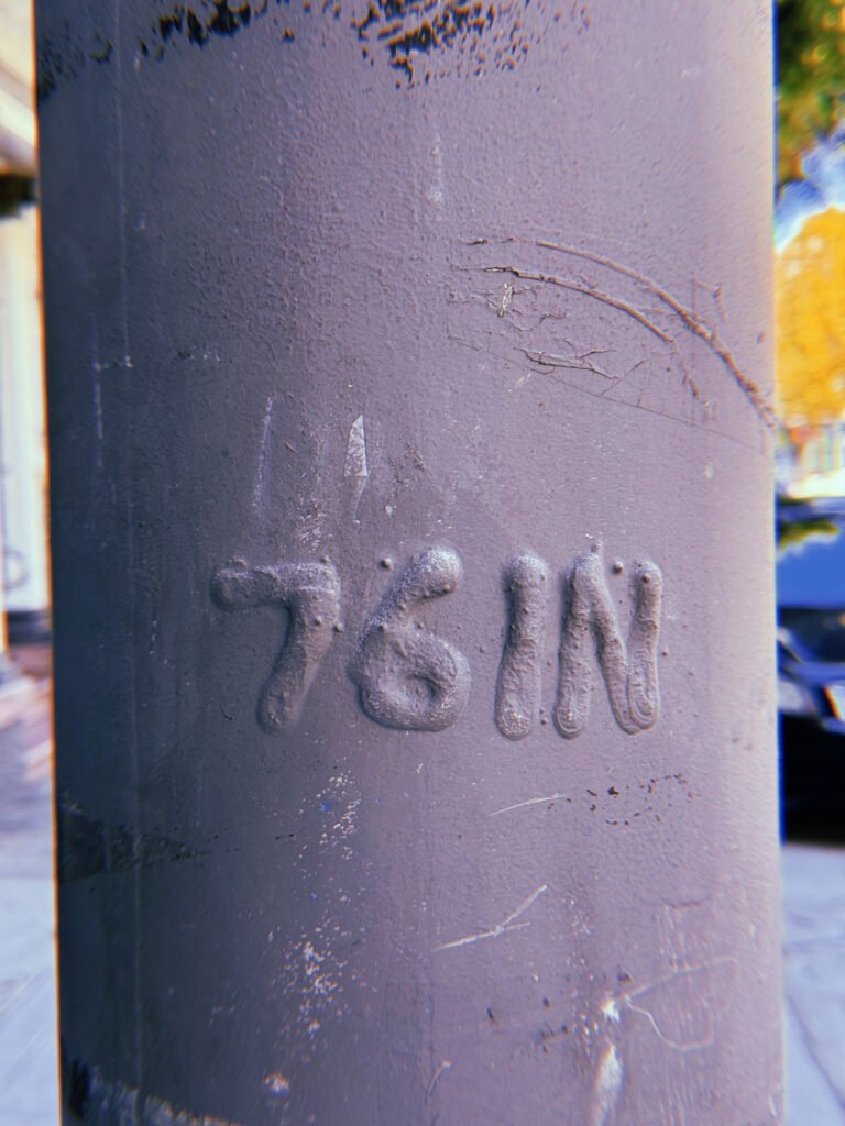 Raised "76 IN" marking embossed on a dark gray-painted metal utility pole in San Francisco. Thick paint covers the lettering, with scratches, cracks, and surface wear visible around it.