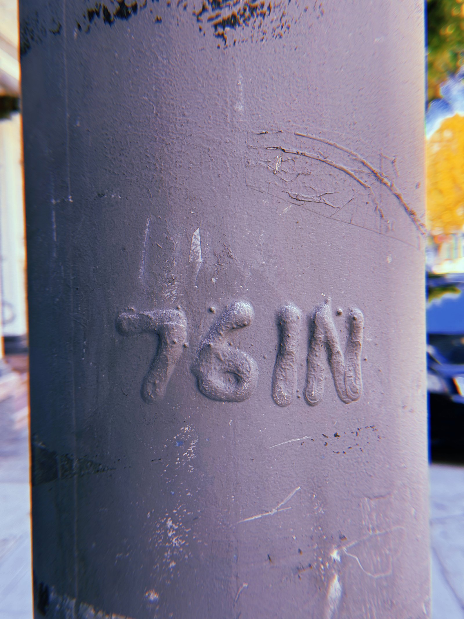 Raised "76 IN" marking embossed on a dark gray-painted metal utility pole in San Francisco. Thick paint covers the lettering, with scratches, cracks, and surface wear visible around it.
