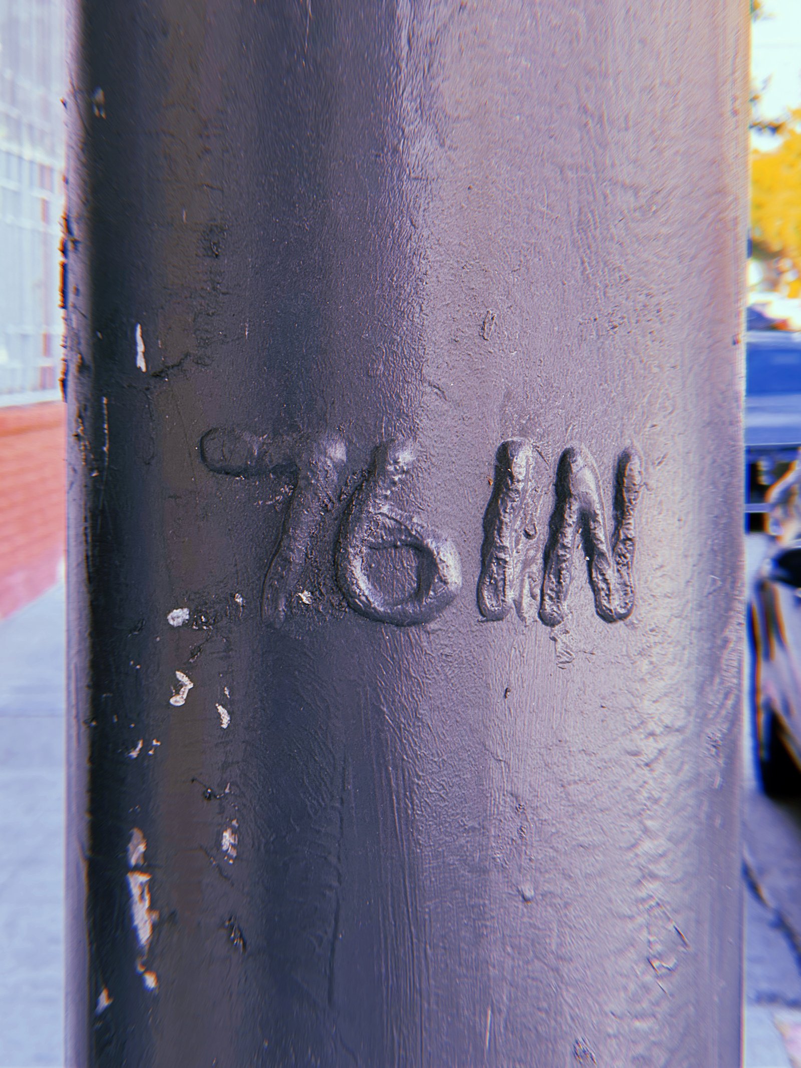 Raised marking "76 IN" on a dark gray-painted metal utility pole in San Francisco. Thick layers of paint partially obscure the lettering, with a city sidewalk and parked vehicles visible in the background.