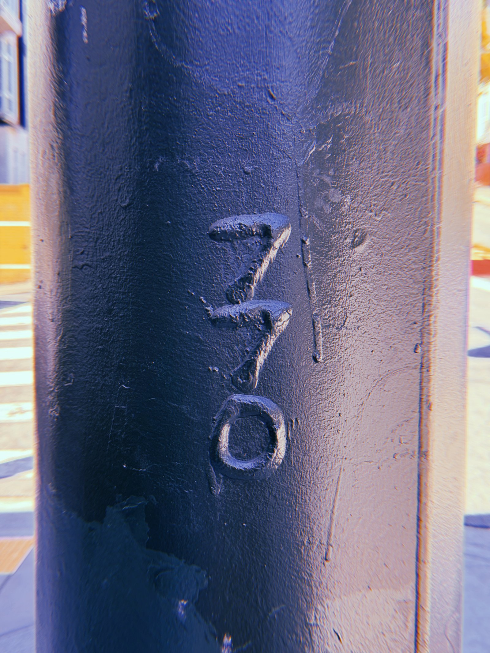 Vertical numbers 770 embossed on a dark blue-painted metal utility pole in San Francisco, with thick layers of paint, scratches, and a street intersection visible in the background.