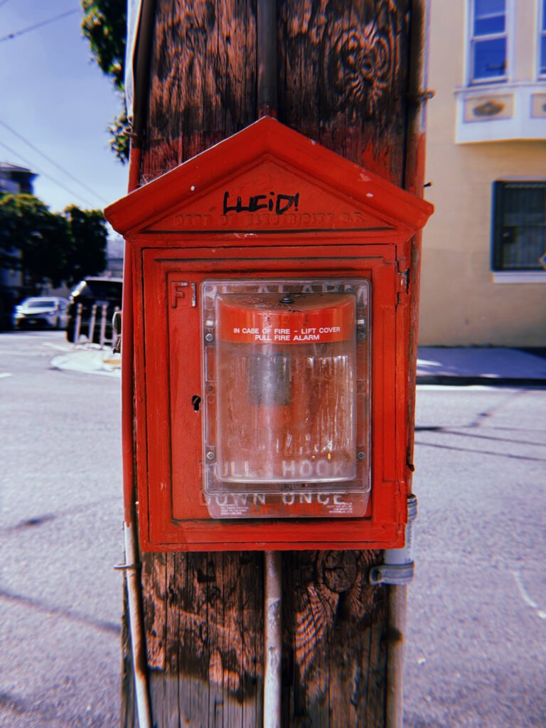 Red cast-iron fire alarm box mounted on a weathered wooden utility pole at a San Francisco street corner. The peaked housing features faded Department of Electricity lettering and black graffiti. A transparent protective cover encloses the alarm mechanism, with a residential building and parked cars in the background.