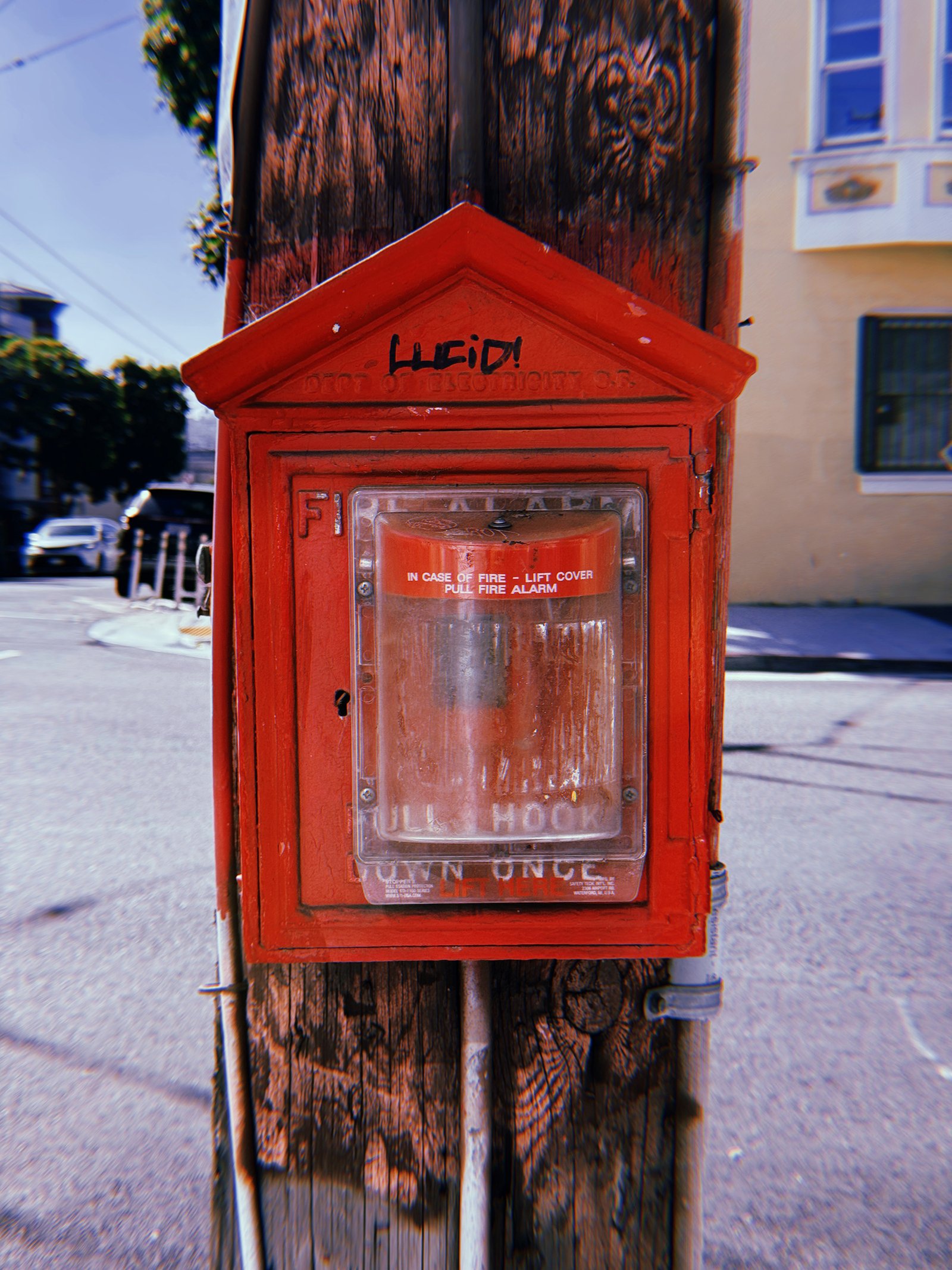 Red cast-iron fire alarm box mounted on a weathered wooden utility pole at a San Francisco street corner. The peaked housing features faded Department of Electricity lettering and black graffiti. A transparent protective cover encloses the alarm mechanism, with a residential building and parked cars in the background.
