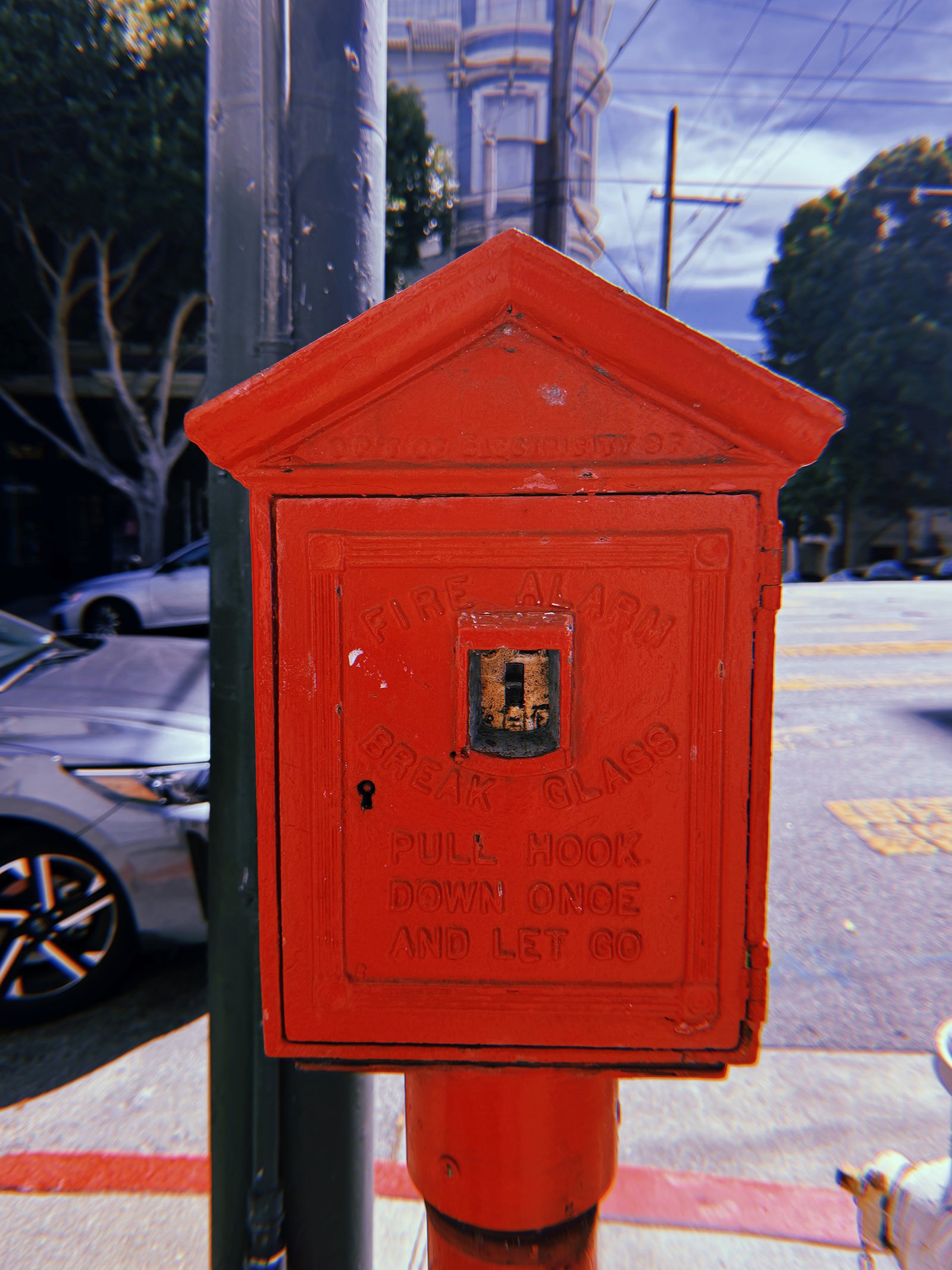 Historic red cast-iron fire alarm box on a San Francisco street corner. The peaked housing features embossed lettering reading “Fire Alarm,” “Break Glass,” and “Pull Hook Down Once and Let Go.” A small exposed metal mechanism sits at the center, with parked cars, utility poles, overhead wires, and residential buildings in the background.