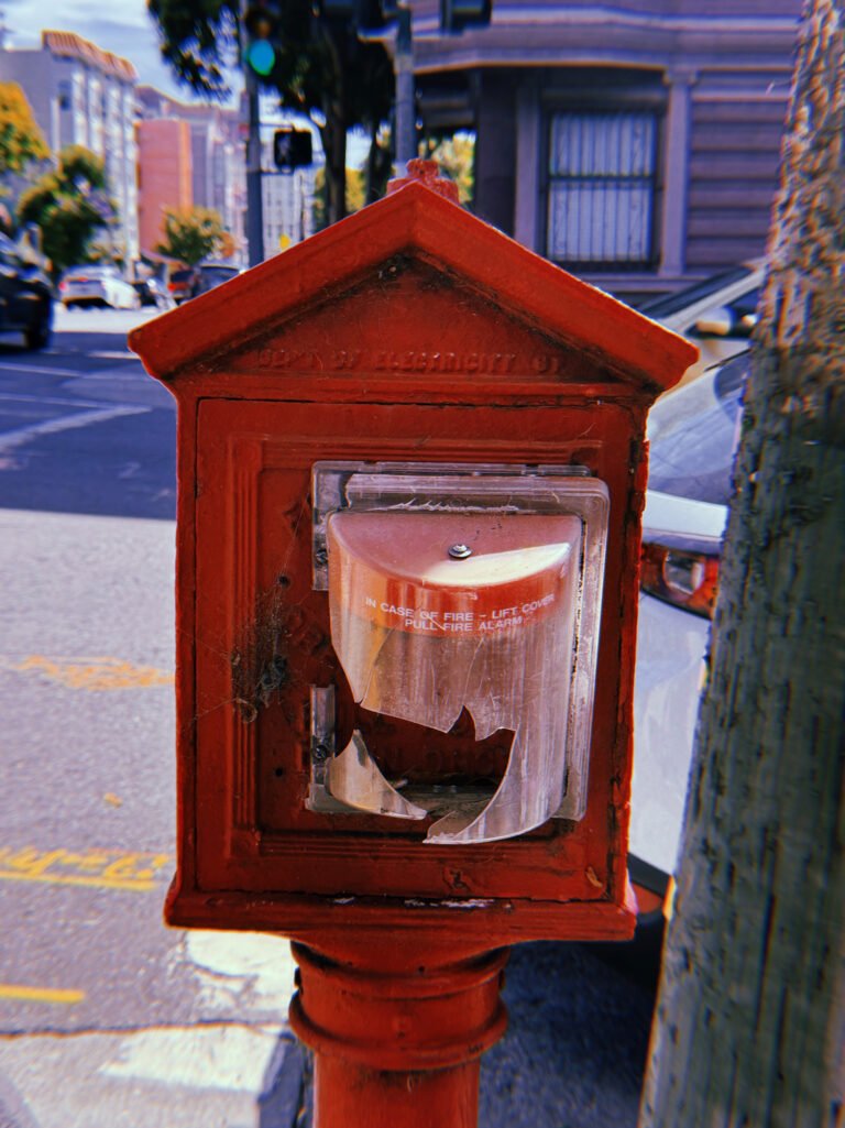 Red cast-iron fire alarm box mounted on a pole at a San Francisco street corner. The historic peaked housing contains a modern fire alarm pull station with a broken transparent protective cover. A tree trunk, parked white vehicle, and residential buildings appear in the background.
