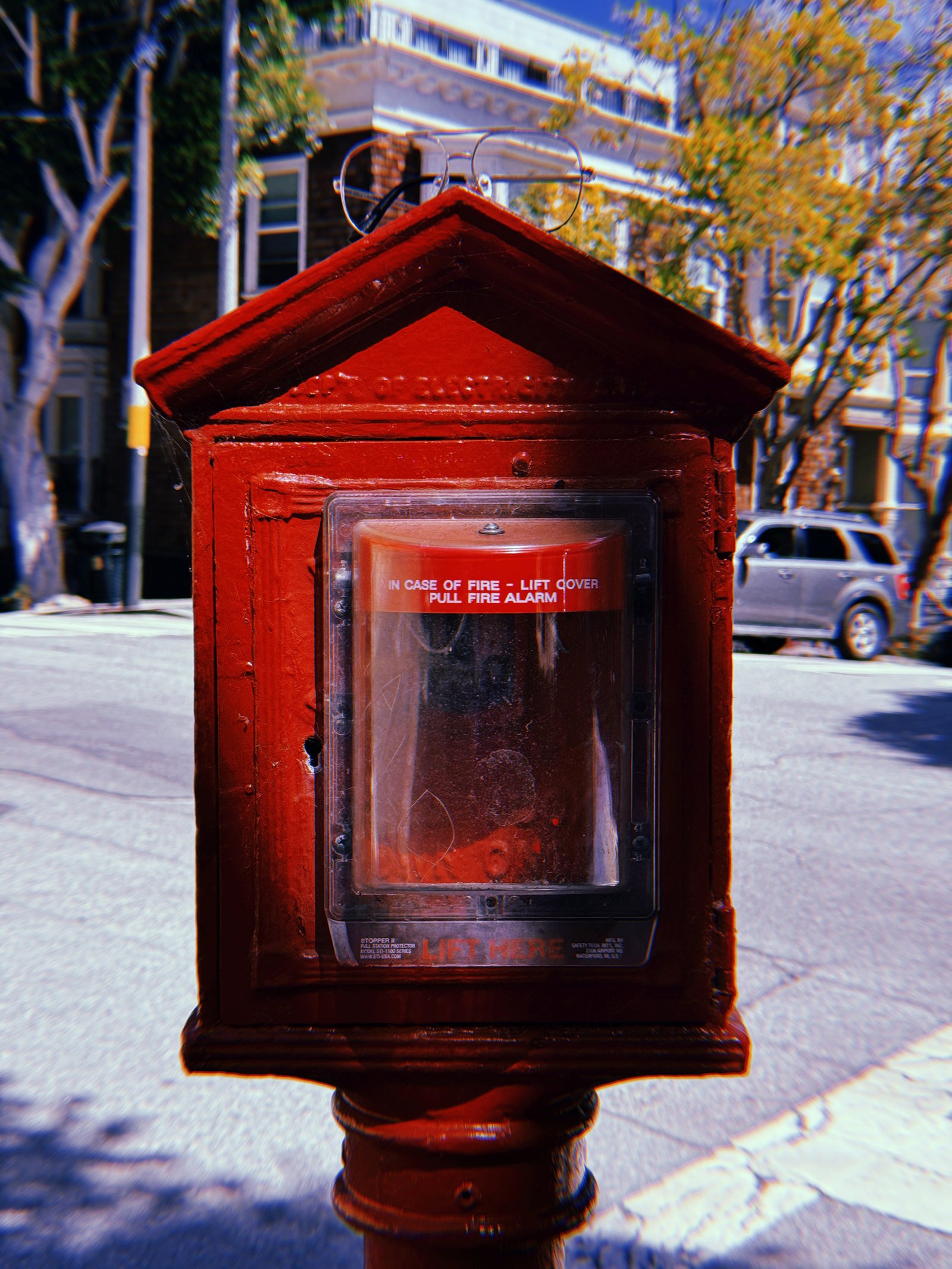 Red cast-iron fire alarm box mounted on a pole at a San Francisco street corner. The historic peaked housing contains a modern fire alarm pull station behind a scratched transparent protective cover. A pair of eyeglasses rests on top of the box, with residential buildings, trees, and a parked SUV in the background.