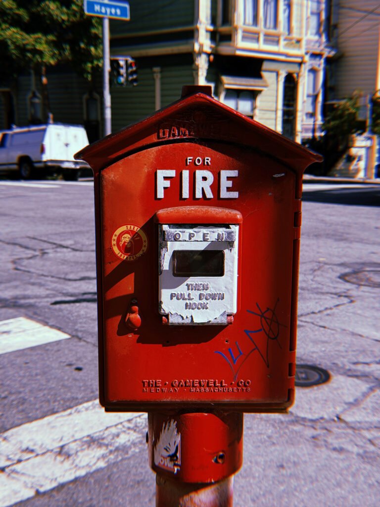 Red Gamewell fire alarm box mounted on a pole at a San Francisco street corner. White “FOR FIRE” lettering appears above a silver pull-down mechanism. Stickers, graffiti, and worn paint cover the metal housing, with Victorian buildings and a street intersection in the background.