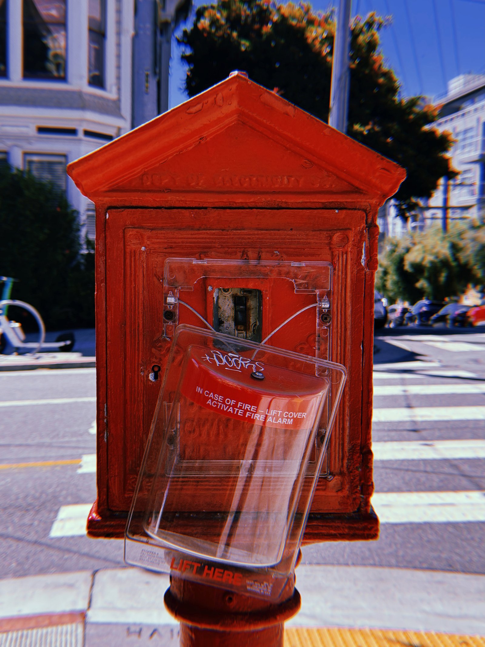 Red cast-iron fire alarm box mounted on a pole at a San Francisco intersection. Its transparent protective cover hangs open at an angle, exposing the recessed alarm mechanism. Victorian buildings, parked cars, and a pedestrian crossing appear in the background.