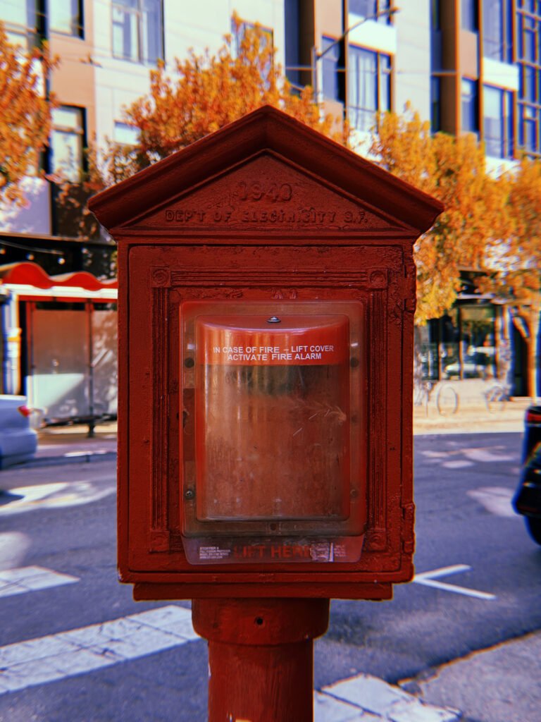 Red cast-iron fire alarm box mounted on a pole at a San Francisco intersection. The historic housing is marked “Dept of Electricity S.F.” and contains a modern fire alarm pull station behind a transparent protective cover. Autumn trees, buildings, and a food trailer appear in the background.