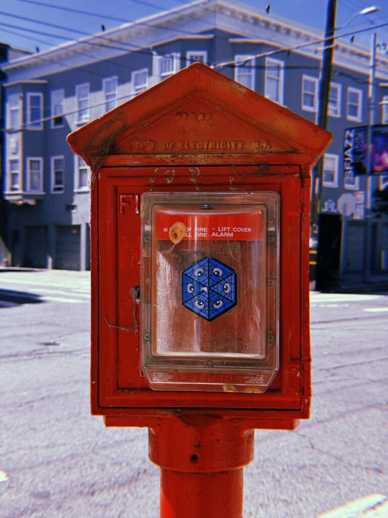 Red fire alarm box mounted on a pole at a San Francisco intersection. The weathered cast-iron housing contains a modern fire alarm pull station behind a transparent protective cover with a blue geometric sticker. Residential buildings and overhead wires appear in the background.