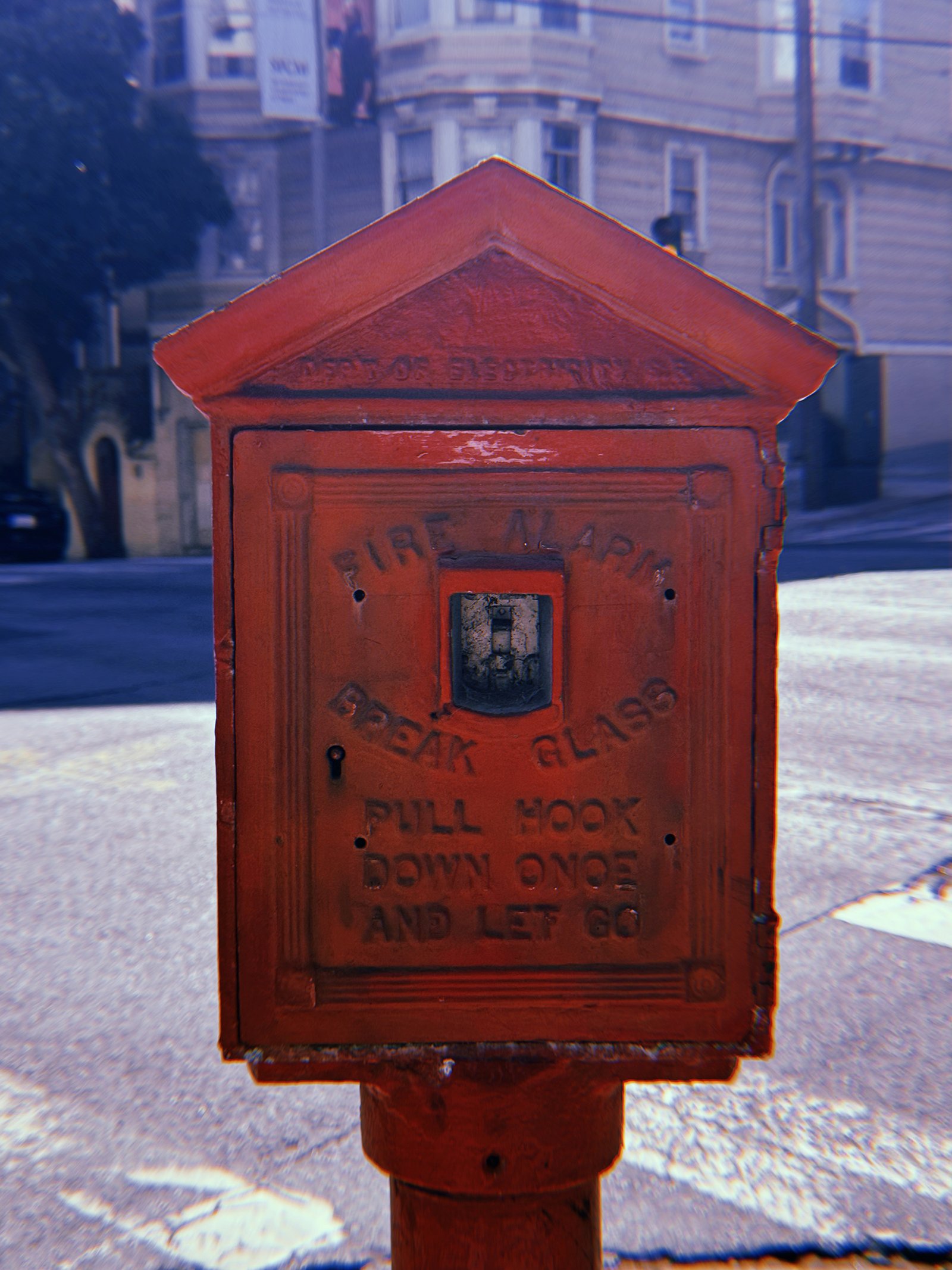 Red cast-iron fire alarm box mounted on a pole at a San Francisco street corner. Weathered lettering reads “FIRE ALARM” and “BREAK GLASS PULL HOOK DOWN ONCE AND LET GO.” Residential buildings and a sloping street appear in the background.