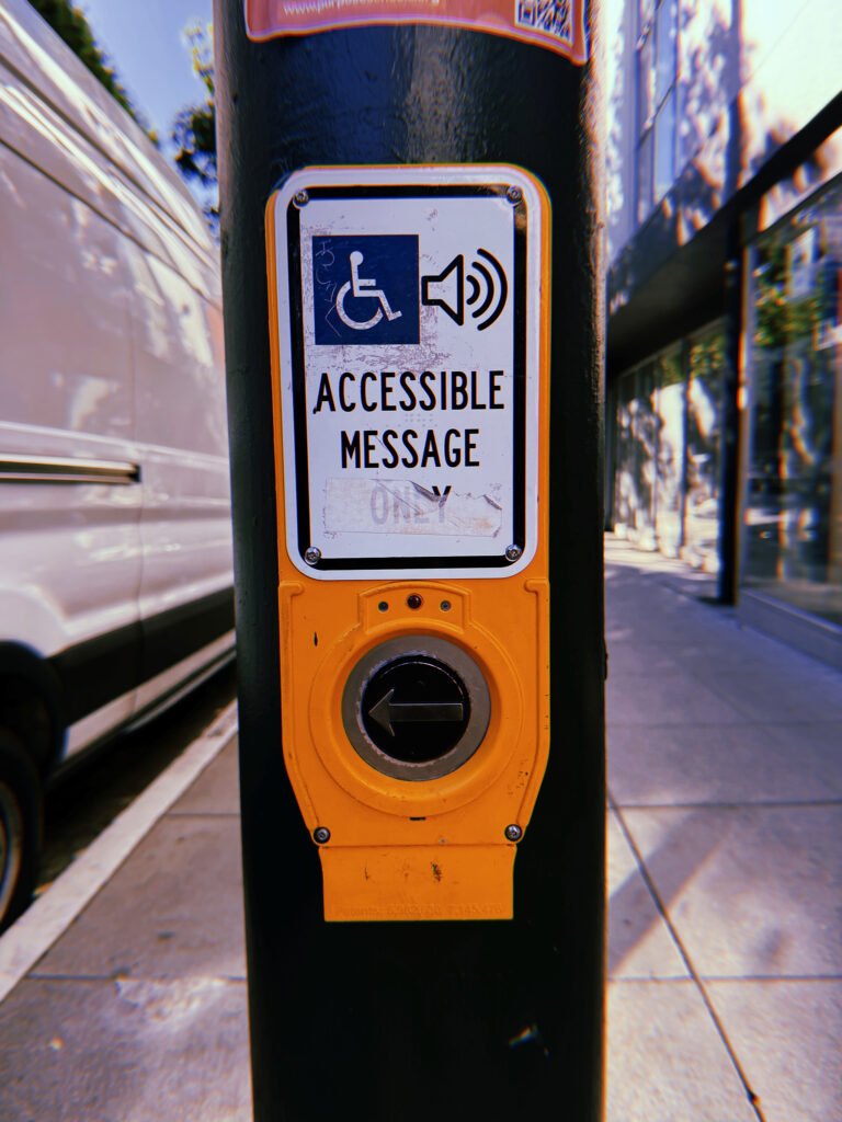 Yellow pedestrian crossing push button mounted on a dark green metal pole in San Francisco. The white sign displays wheelchair accessibility and speaker symbols above the words “Accessible Message Only,” with the word “Only” partially covered by a faded sticker. A circular black button with a left-pointing arrow sits below. A white van and urban storefronts appear in the background.