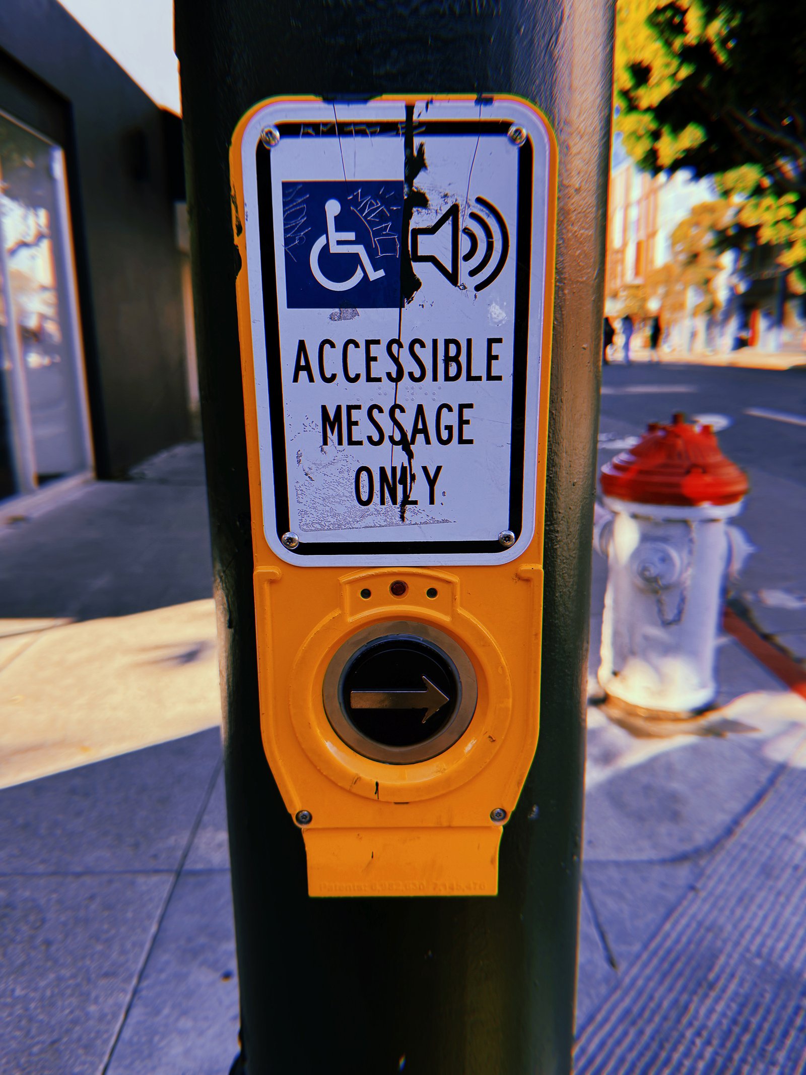 Yellow pedestrian crossing push button mounted on a dark green pole in San Francisco. The weathered sign reads “Accessible Message Only” with wheelchair and speaker symbols, partially obscured by black paint and scratches. A circular black button with a right-pointing arrow sits below. A white fire hydrant with a red top is visible in the background.