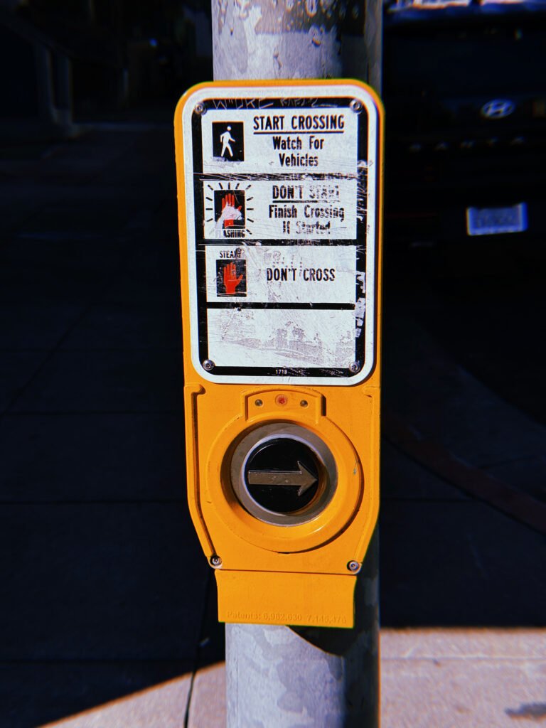 Close-up of a yellow pedestrian crossing push button mounted on a gray metal pole in San Francisco. The weathered instruction panel displays walking and red hand symbols with directions for crossing. A circular black button with a right-pointing arrow sits below the scratched sign, against a dark sidewalk background.
