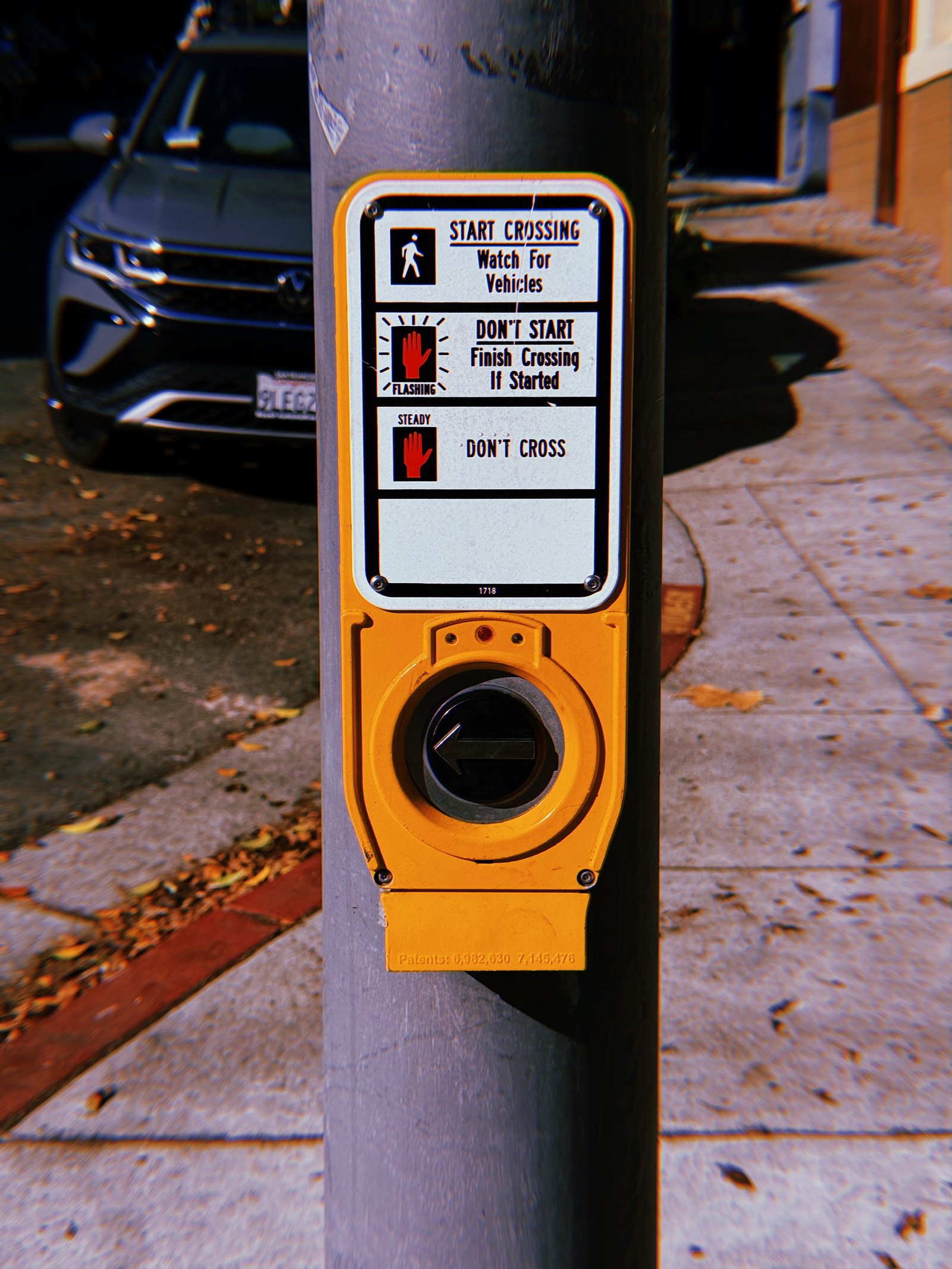 Yellow pedestrian crossing push button mounted on a gray metal pole along a San Francisco sidewalk. A rectangular instruction sign above displays walking and red hand symbols with directions for crossing safely. The circular button features a left-pointing arrow, with a parked SUV, fallen leaves, and a sunlit sidewalk in the background.