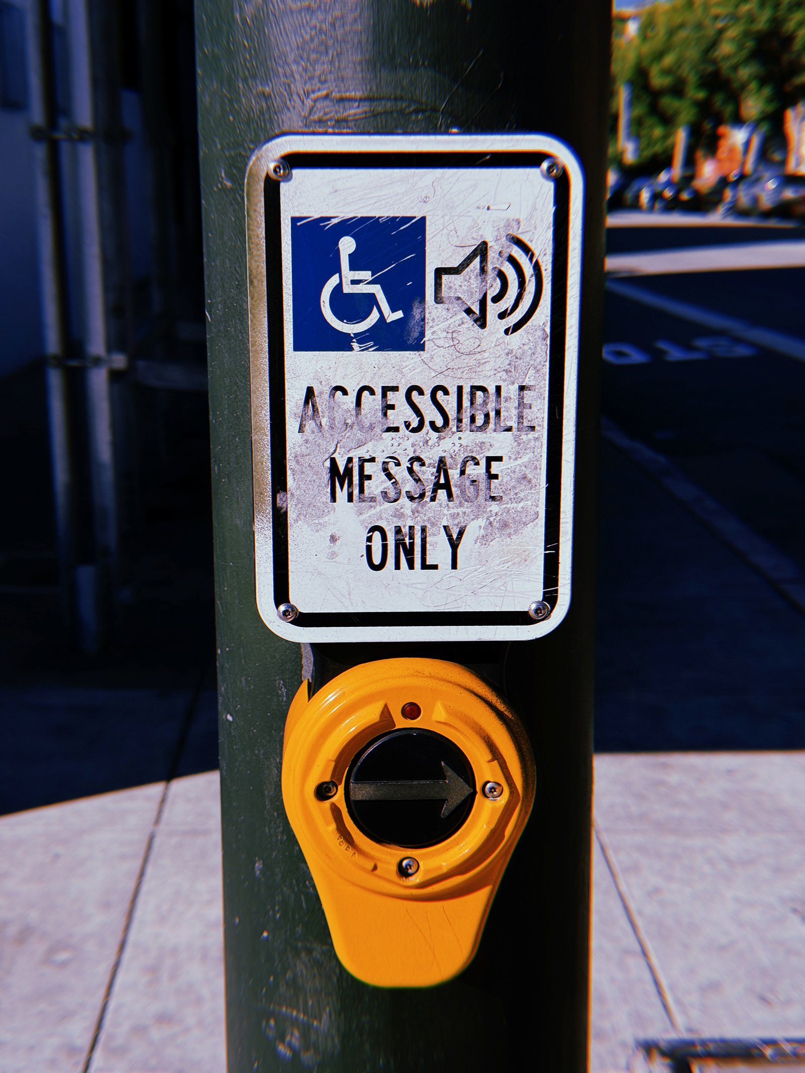 Yellow pedestrian crossing push button mounted on a dark green metal pole at a San Francisco intersection. A heavily scratched white sign above reads “Accessible Message Only” beneath wheelchair and speaker symbols. The circular button features a right-pointing arrow, with a shaded street and sidewalk in the background.