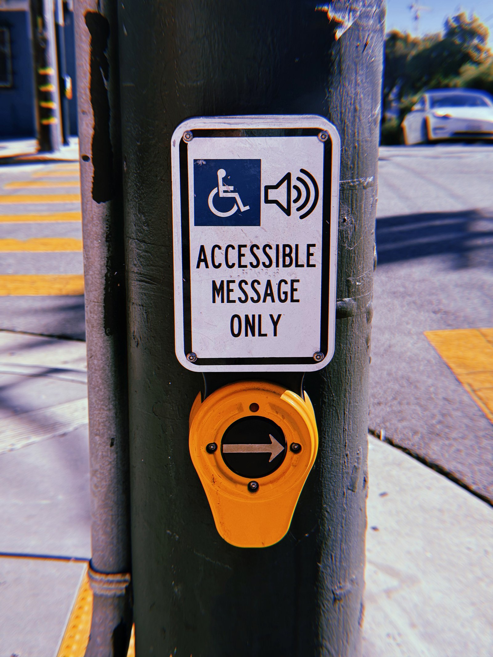 Yellow pedestrian crossing push button mounted on a dark green metal pole at a San Francisco intersection. A white sign above reads “Accessible Message Only” beneath wheelchair and speaker symbols. The circular button features a right-pointing arrow, with yellow crosswalk markings, a curb ramp, and a white car in the background.
