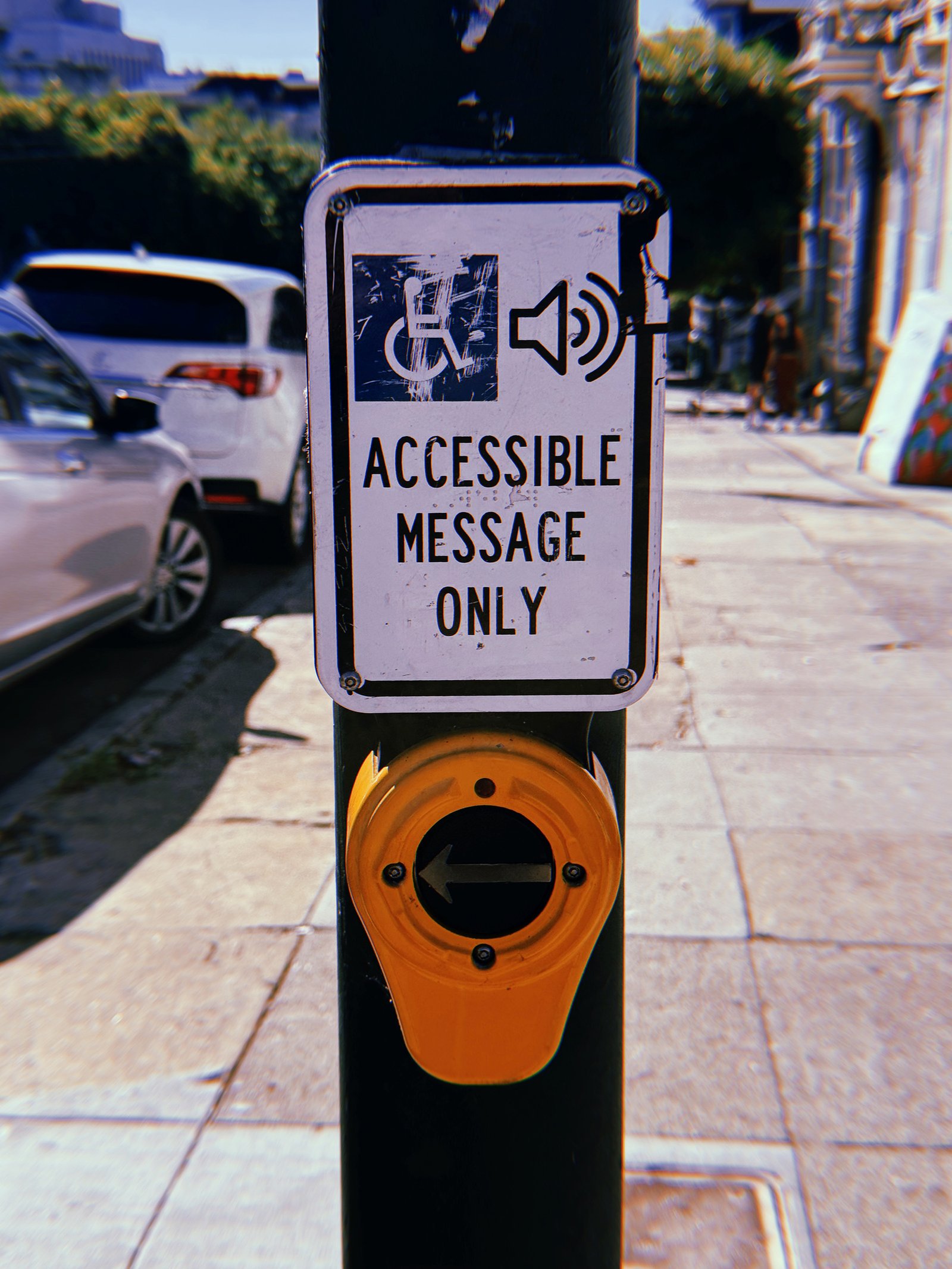 Yellow pedestrian crossing push button mounted on a black metal pole along a San Francisco sidewalk. A scratched white sign above reads “Accessible Message Only” beneath wheelchair and speaker symbols. The circular button features a left-pointing arrow, with parked cars, residential buildings, and a sunlit sidewalk in the background.