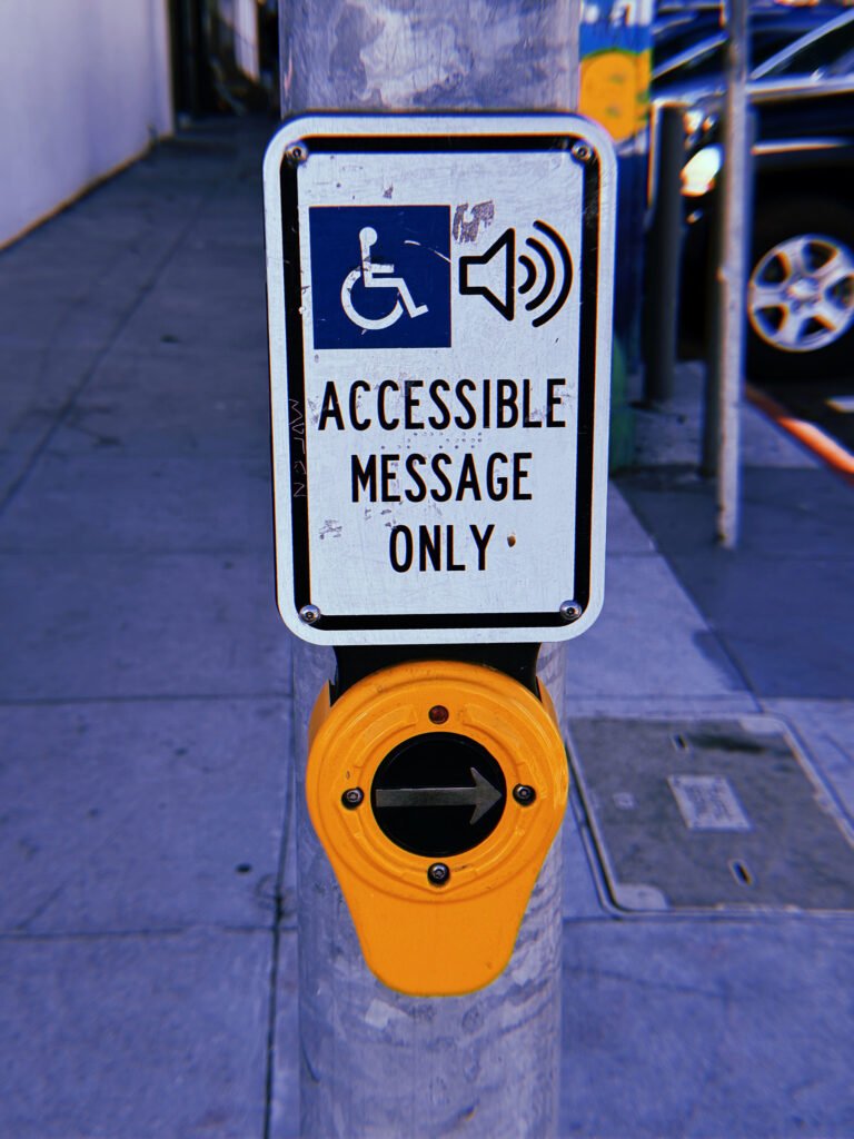 Yellow pedestrian crossing push button mounted on a weathered gray metal pole at a San Francisco intersection. A white rectangular sign above displays wheelchair and speaker symbols with the text “Accessible Message Only.” The circular button features a right-pointing arrow, with a sidewalk, utility covers, and parked vehicles in the background.