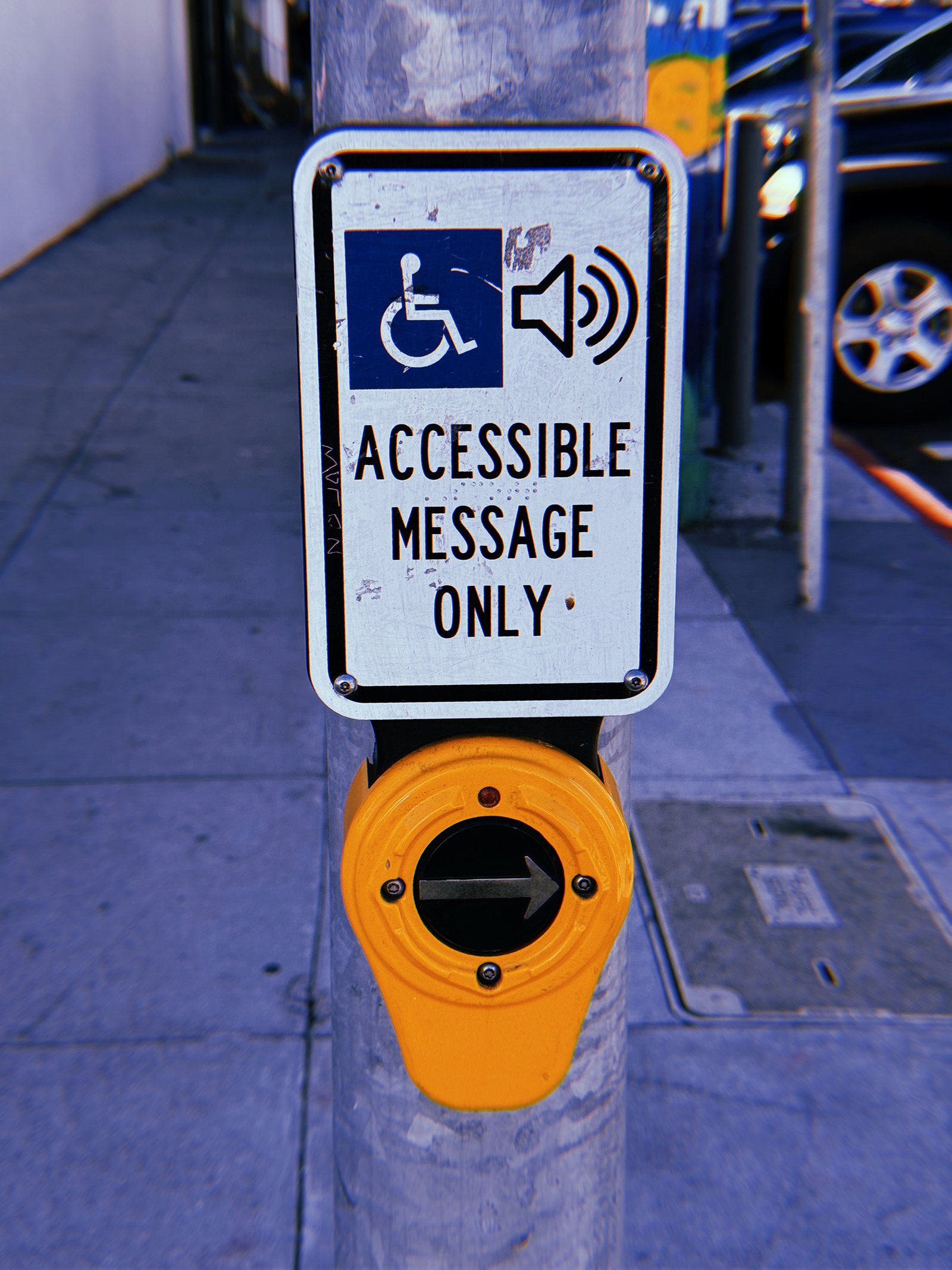 Yellow pedestrian crossing push button mounted on a weathered gray metal pole at a San Francisco intersection. A white rectangular sign above displays wheelchair and speaker symbols with the text “Accessible Message Only.” The circular button features a right-pointing arrow, with a sidewalk, utility covers, and parked vehicles in the background.