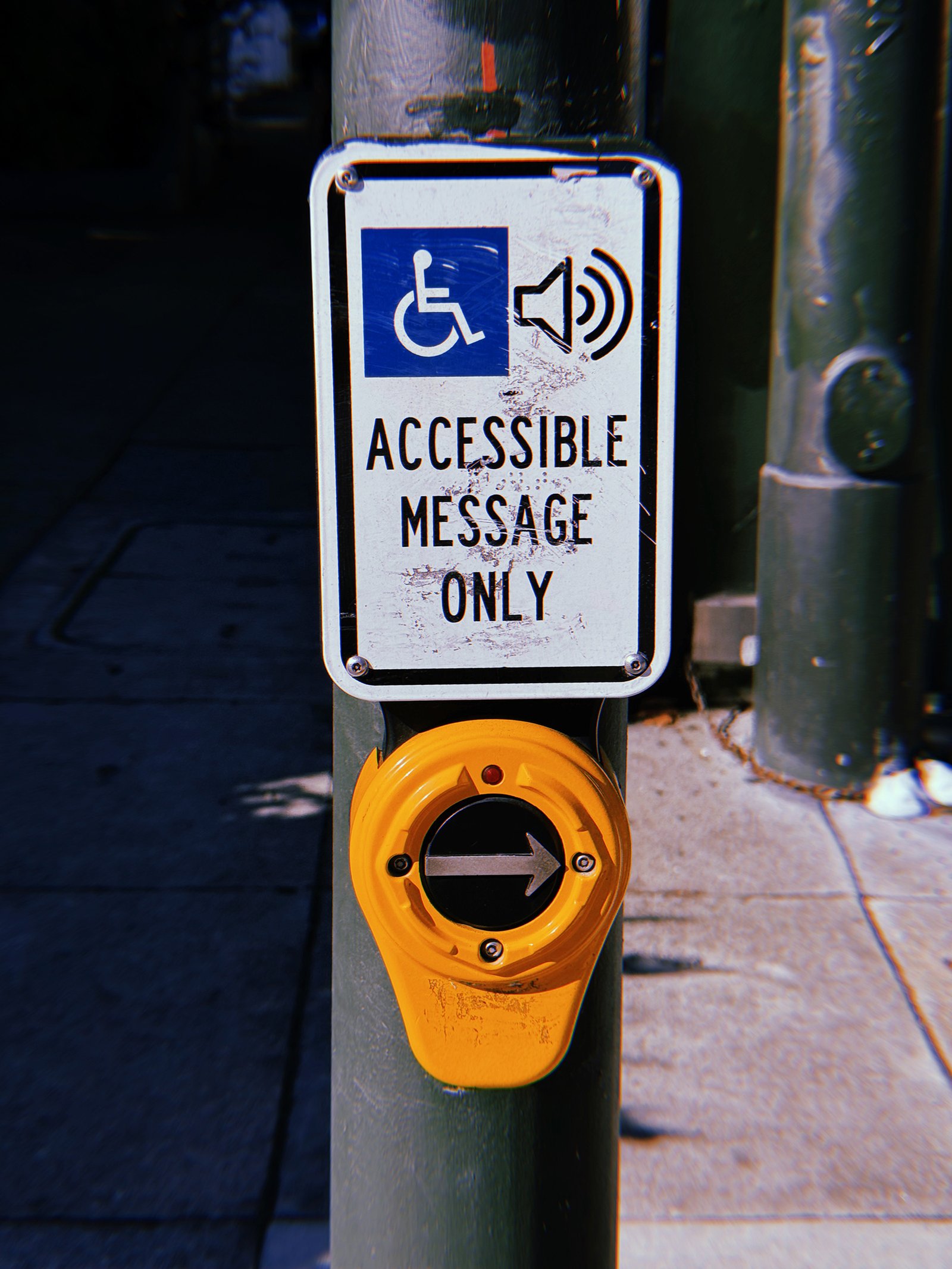 Yellow pedestrian crossing push button mounted on a green metal pole at a San Francisco intersection. Above the button, a weathered white sign reads “Accessible Message Only” beneath wheelchair and speaker symbols. The circular button features a right-pointing arrow, with a shaded sidewalk and traffic signal infrastructure in the background.